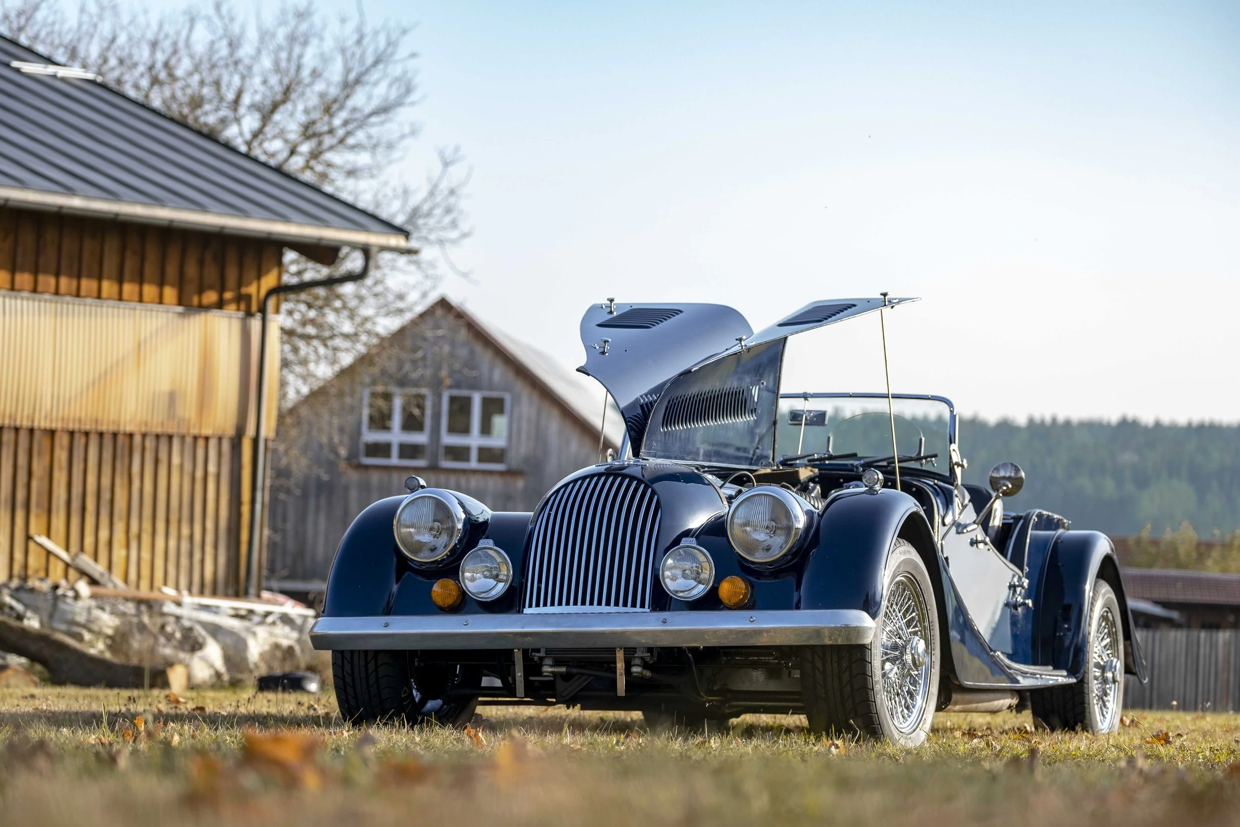 Klassisches Auto mit geöffneter Motorhaube vor rustikalem Gebäude auf einer Wiese.