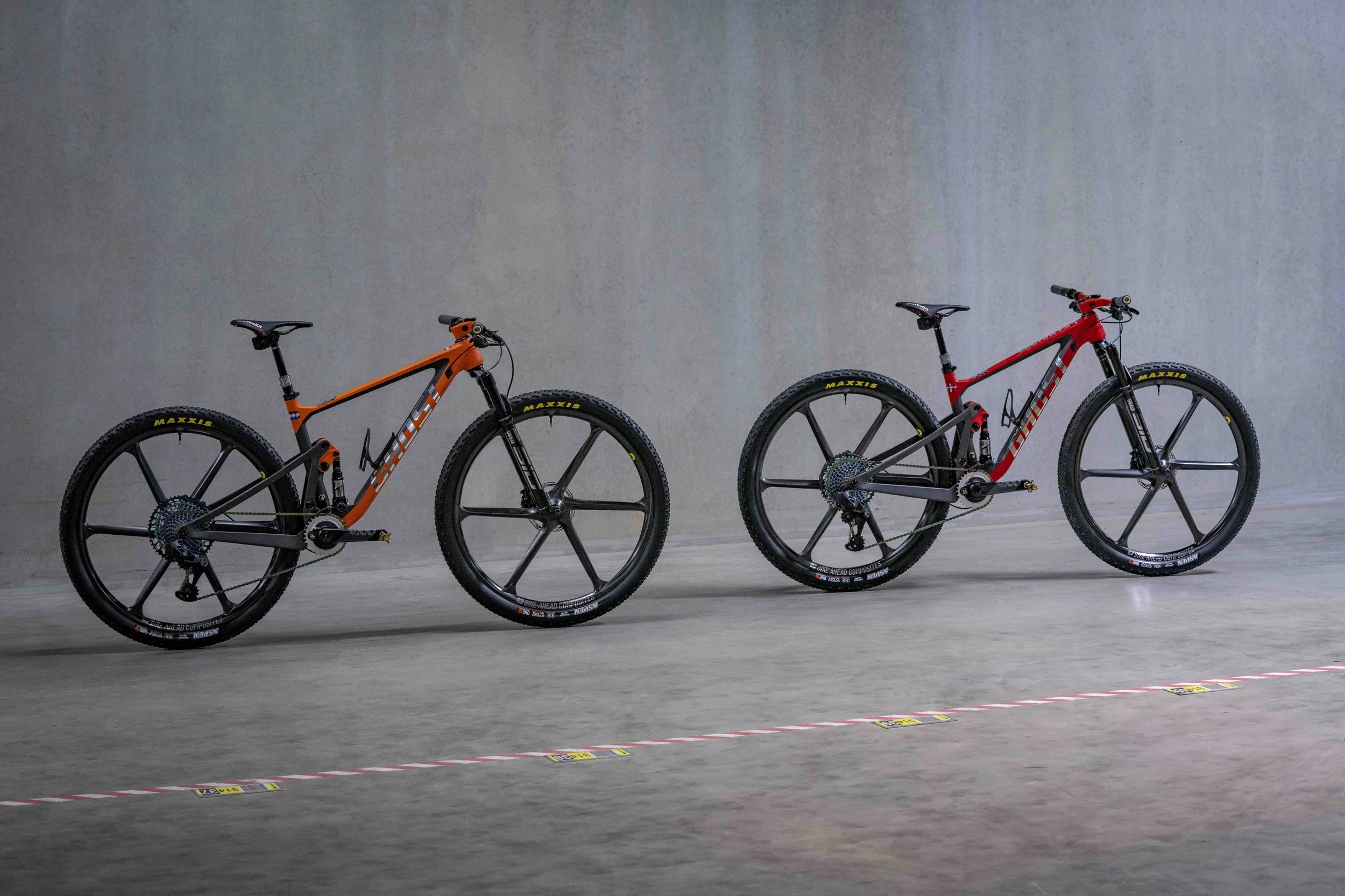 Zwei Mountainbikes, eines orange und eines rot, in einem grauen Innenraum aufgestellt.