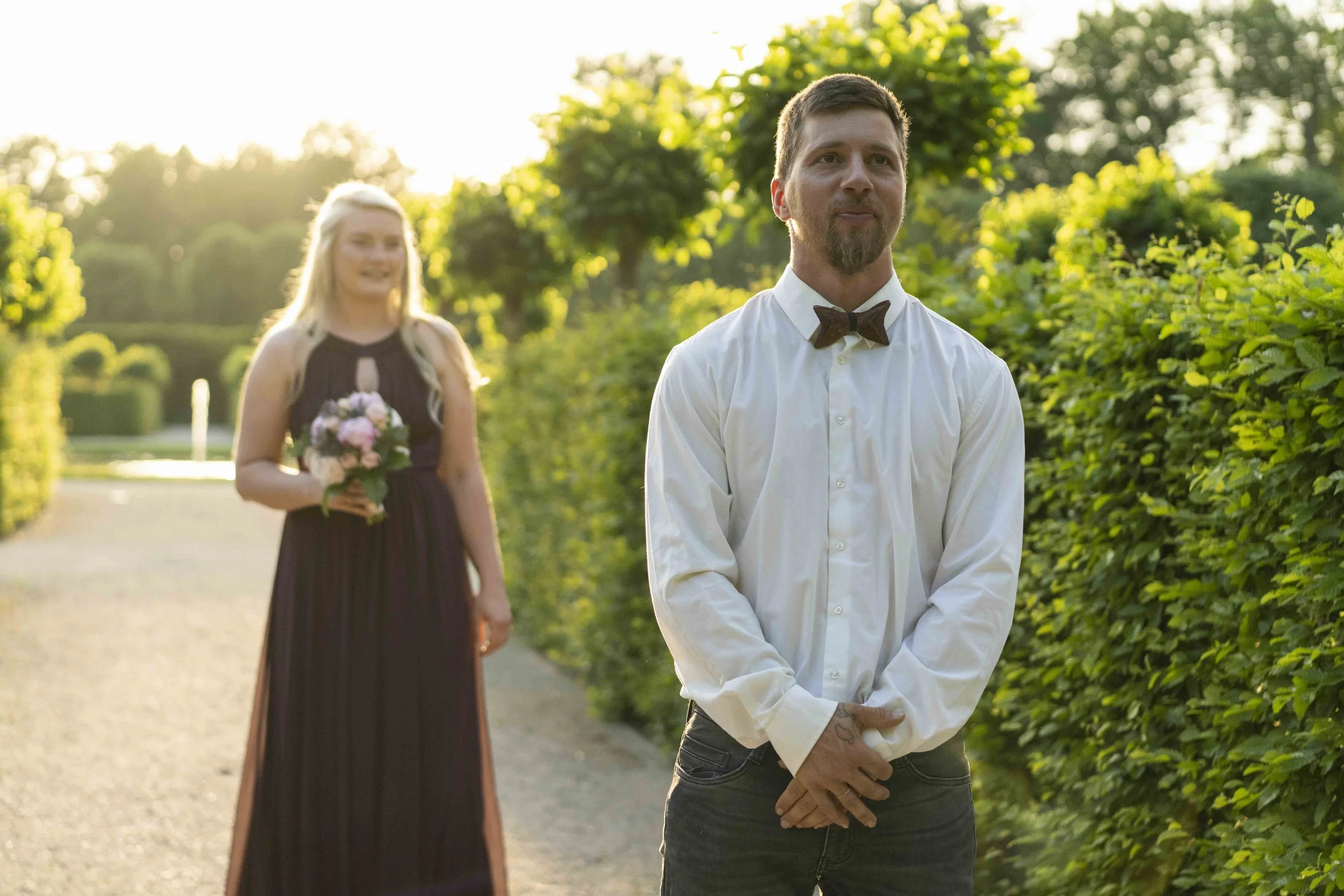 Ein Mann in einem weißen Hemd mit Fliege steht in einem Gartenweg. Eine Frau in einem langen Kleid hält einen Blumenstrauß und steht hinter ihm. Die Szene ist von grünem Laub umgeben, und die Sonne scheint im Hintergrund.