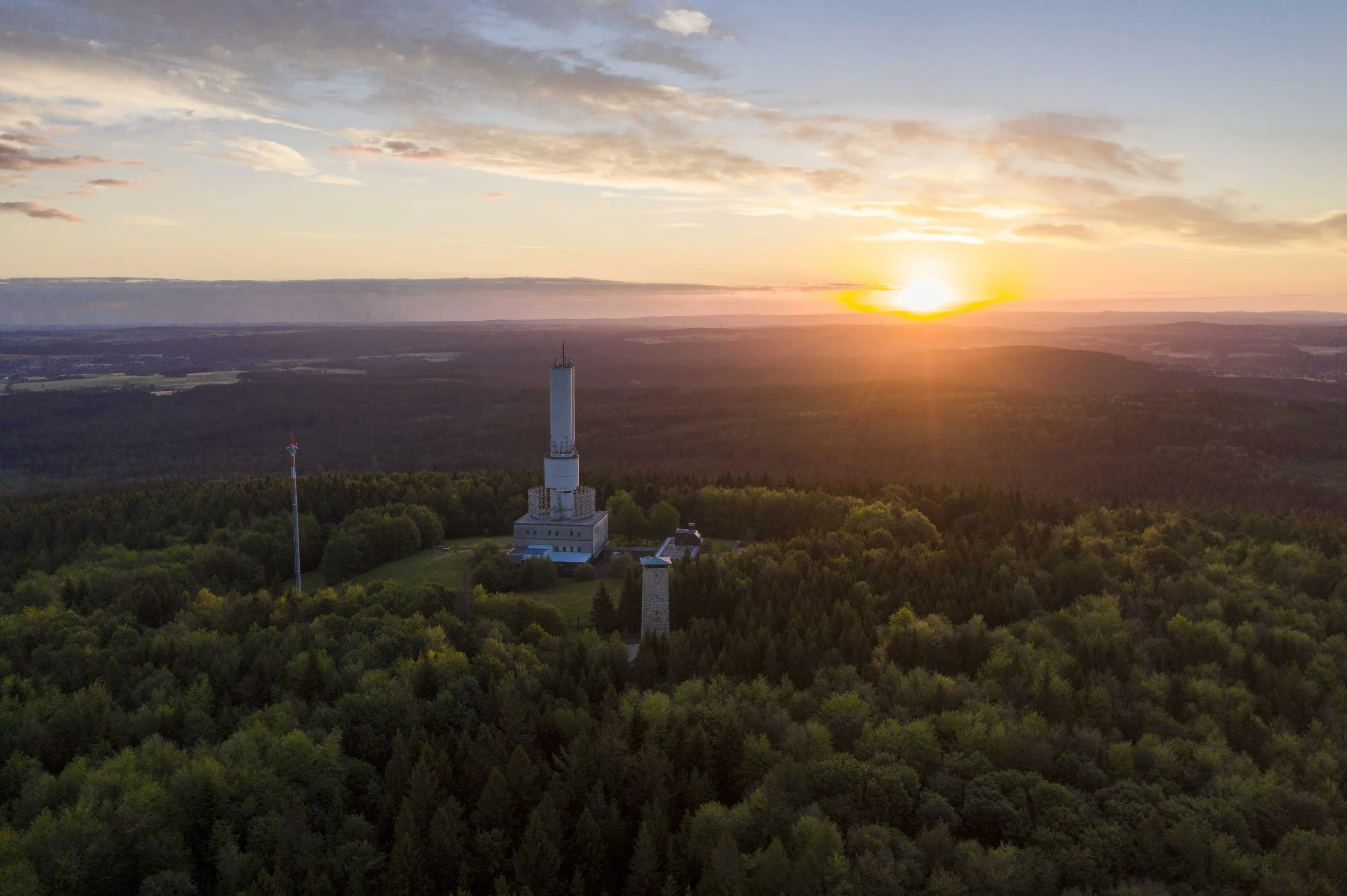 Ein Turm und ein Sendemast in einem Waldgebiet bei Sonnenuntergang.