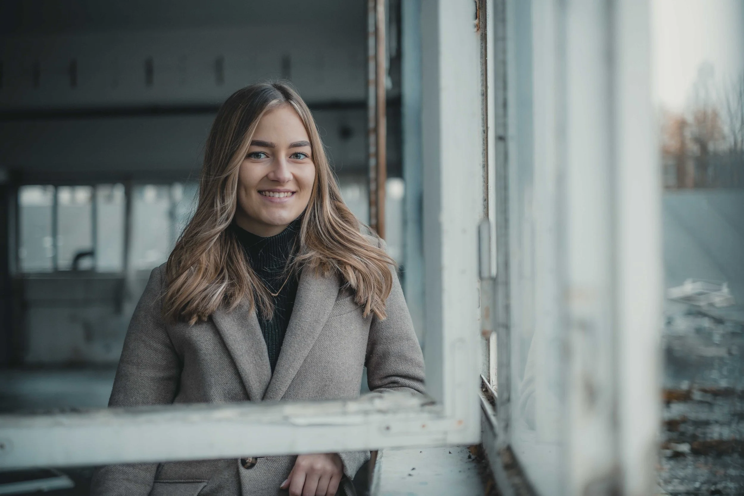 Junge Frau in grauem Mantel lächelt durch ein offenes Fenster, im Hintergrund unscharfe, industrielle Umgebung.