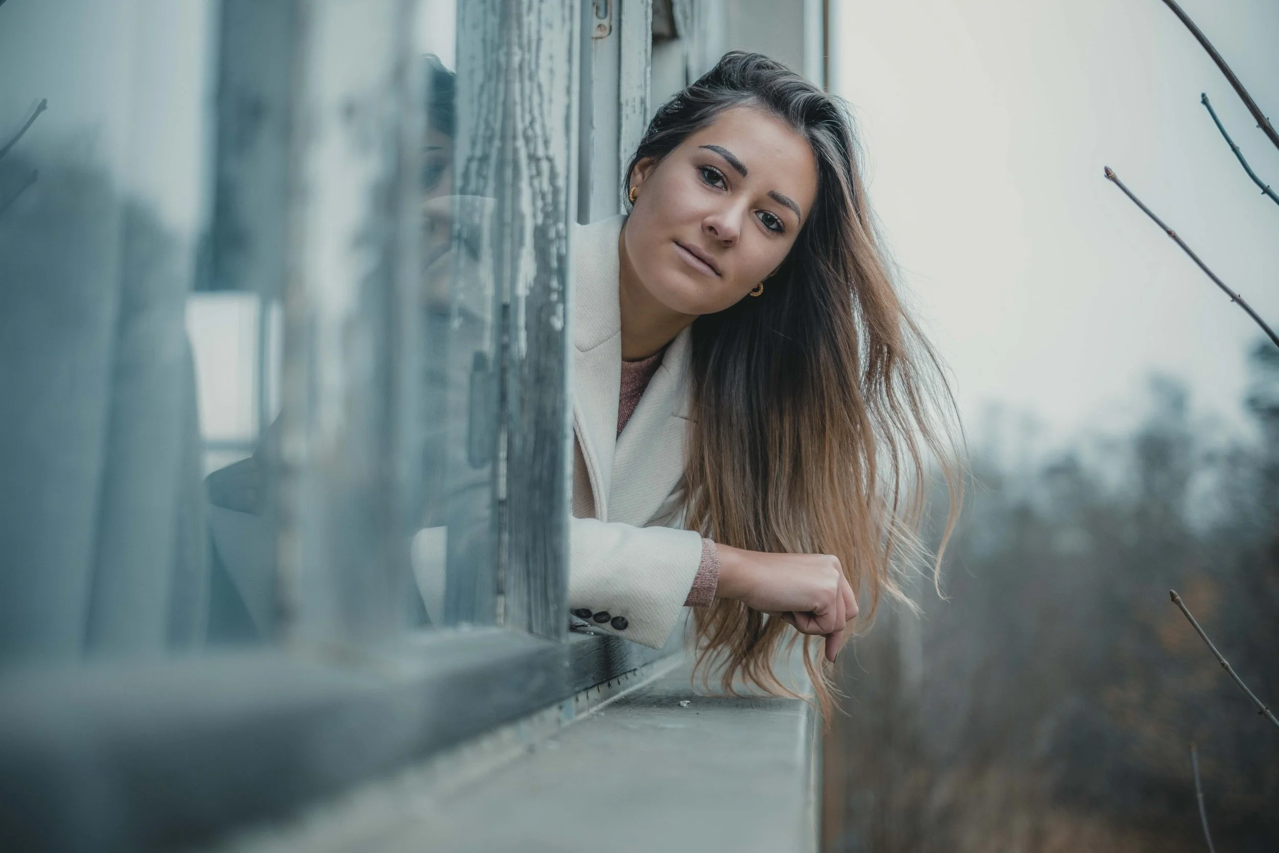 Frau lehnt sich aus einem Fenster, trägt einen beigen Mantel, Hintergrund bewaldete Landschaft.
