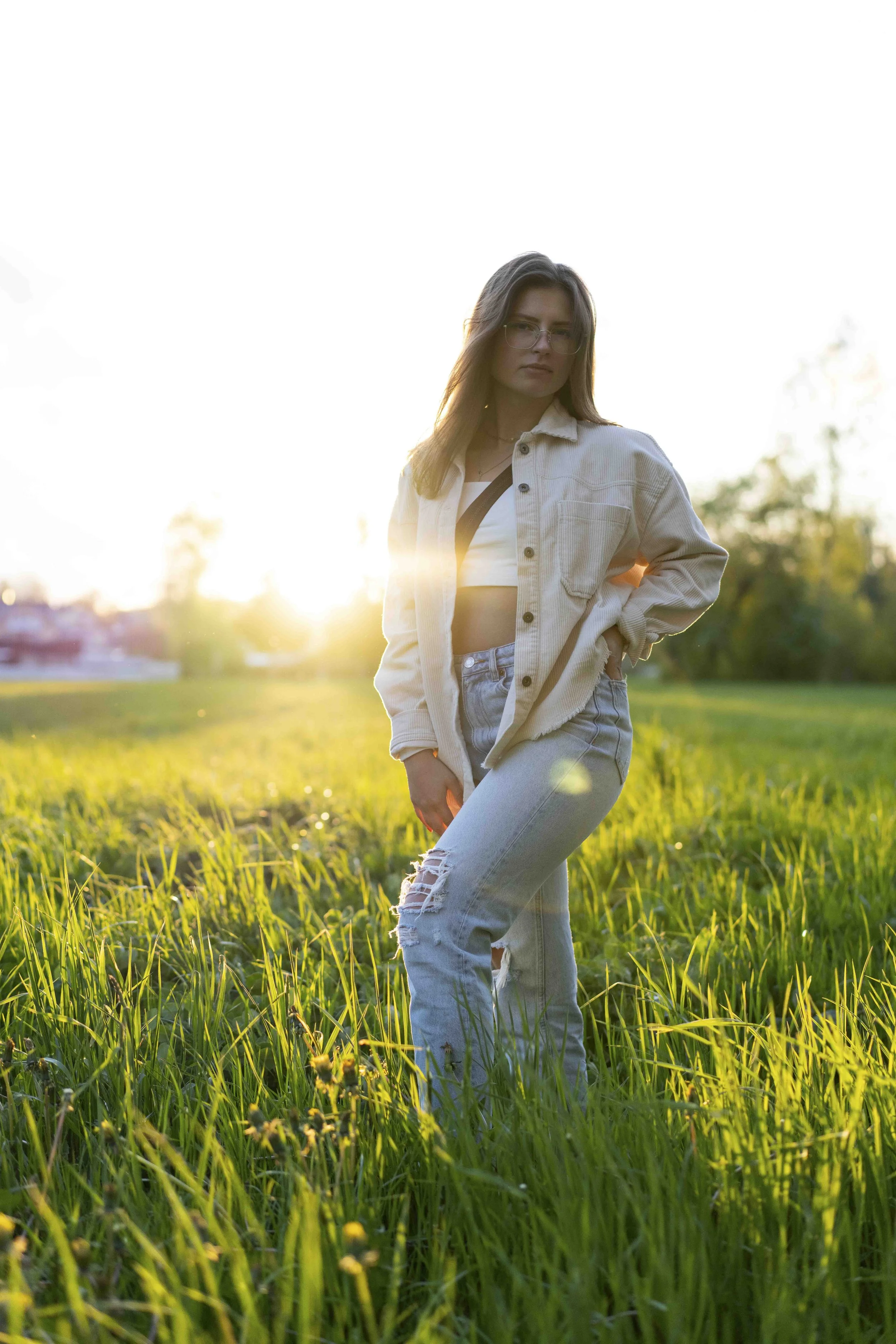 Eine Frau steht im grünen Grasfeld mit Sonnenuntergang im Hintergrund, trägt eine helle Jacke und Jeans.