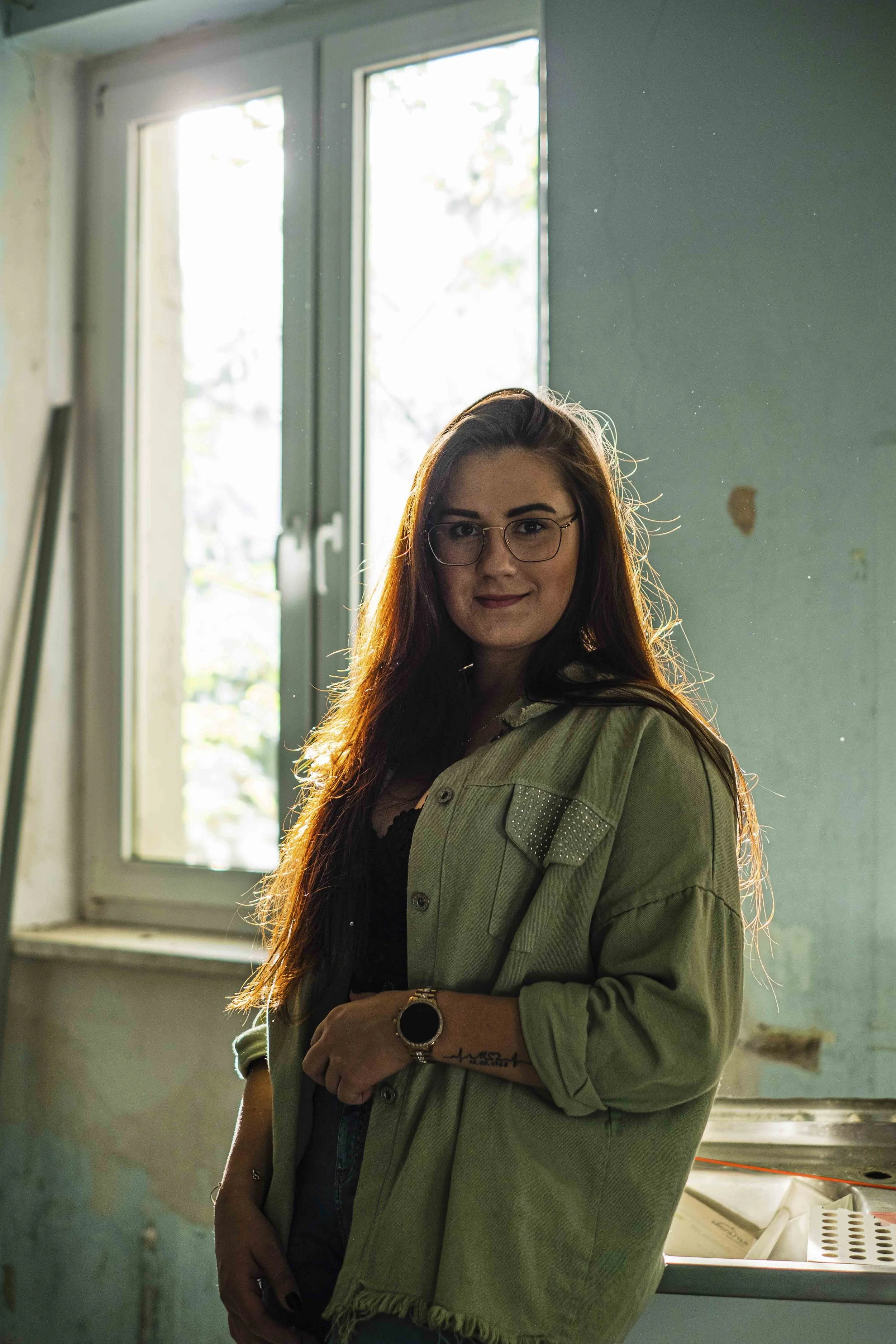 Junge Frau mit langen Haaren und Brille steht in einem Raum mit grün gestrichenen Wänden vor einem Fenster, das Licht hereinlässt. Sie trägt eine grüne Jacke und eine Uhr, und lächelt leicht.