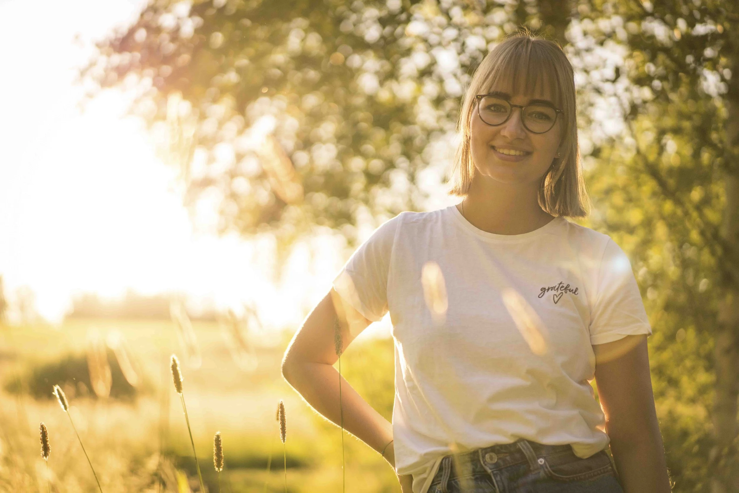 Frau mit Brille steht in einem Feld bei Sonnenuntergang, trägt ein weißes T-Shirt mit der Aufschrift 'grateful'.