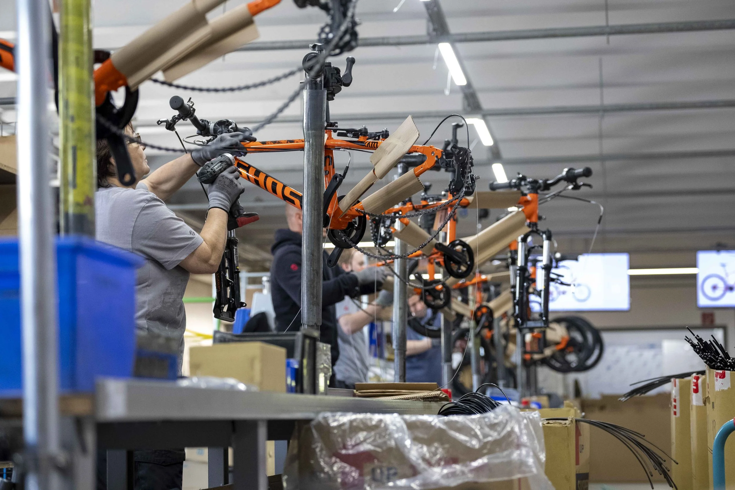 Arbeiter montieren Fahrräder in einer Fabrikhalle, mehrere orange Fahrräder auf einer Produktionslinie, Kartons und Werkzeuge im Vordergrund.