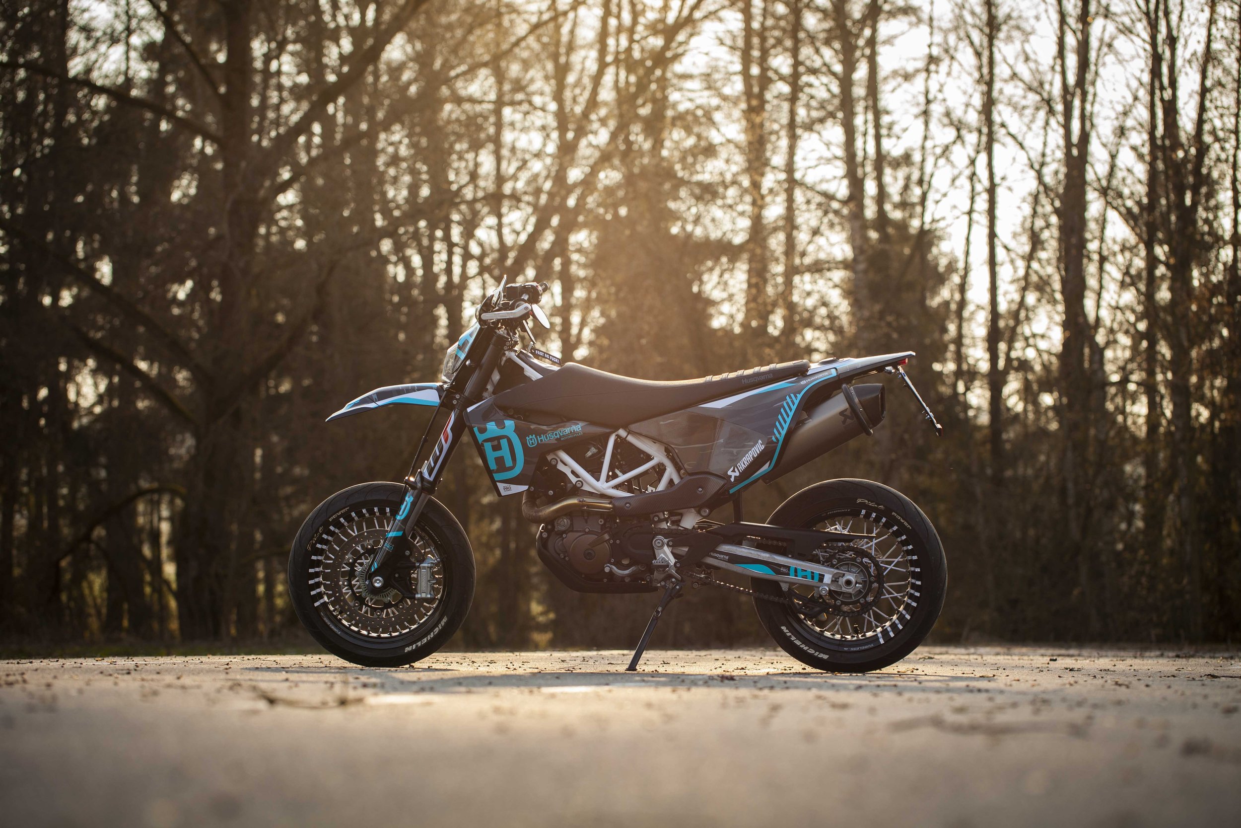 Ein Motorrad steht im Freien auf einer asphaltierten Fläche, im Hintergrund sind Bäume zu sehen. Es ist ein Sportmotorrad, vermutlich eine Supermoto, mit einem schlanken und sportlichen Design in Blau- und Schwarztönen. Die Umgebung wirkt ruhig mit e