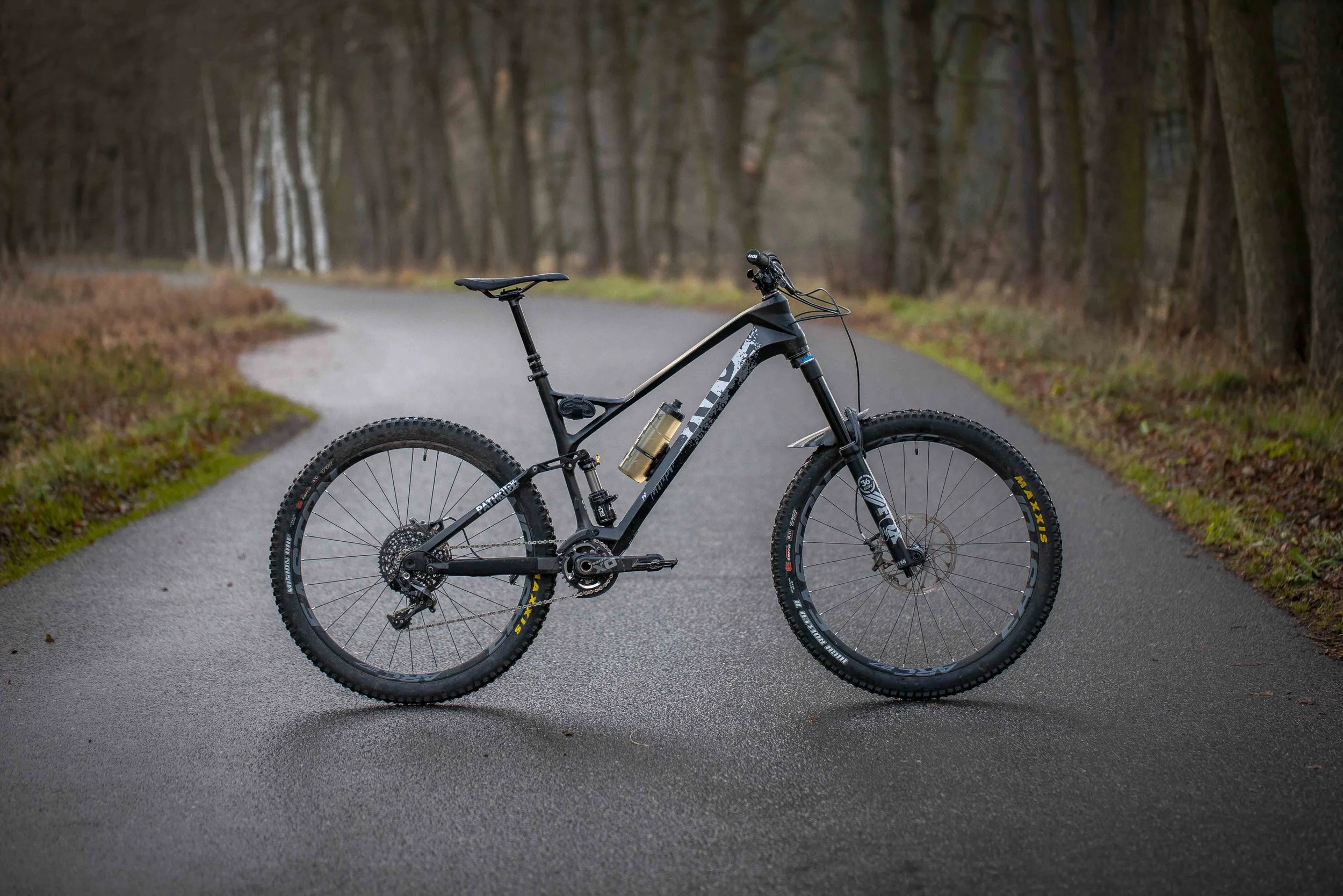 Schwarzes Mountainbike mit dicken Reifen auf einem Waldweg, umgeben von Bäumen.