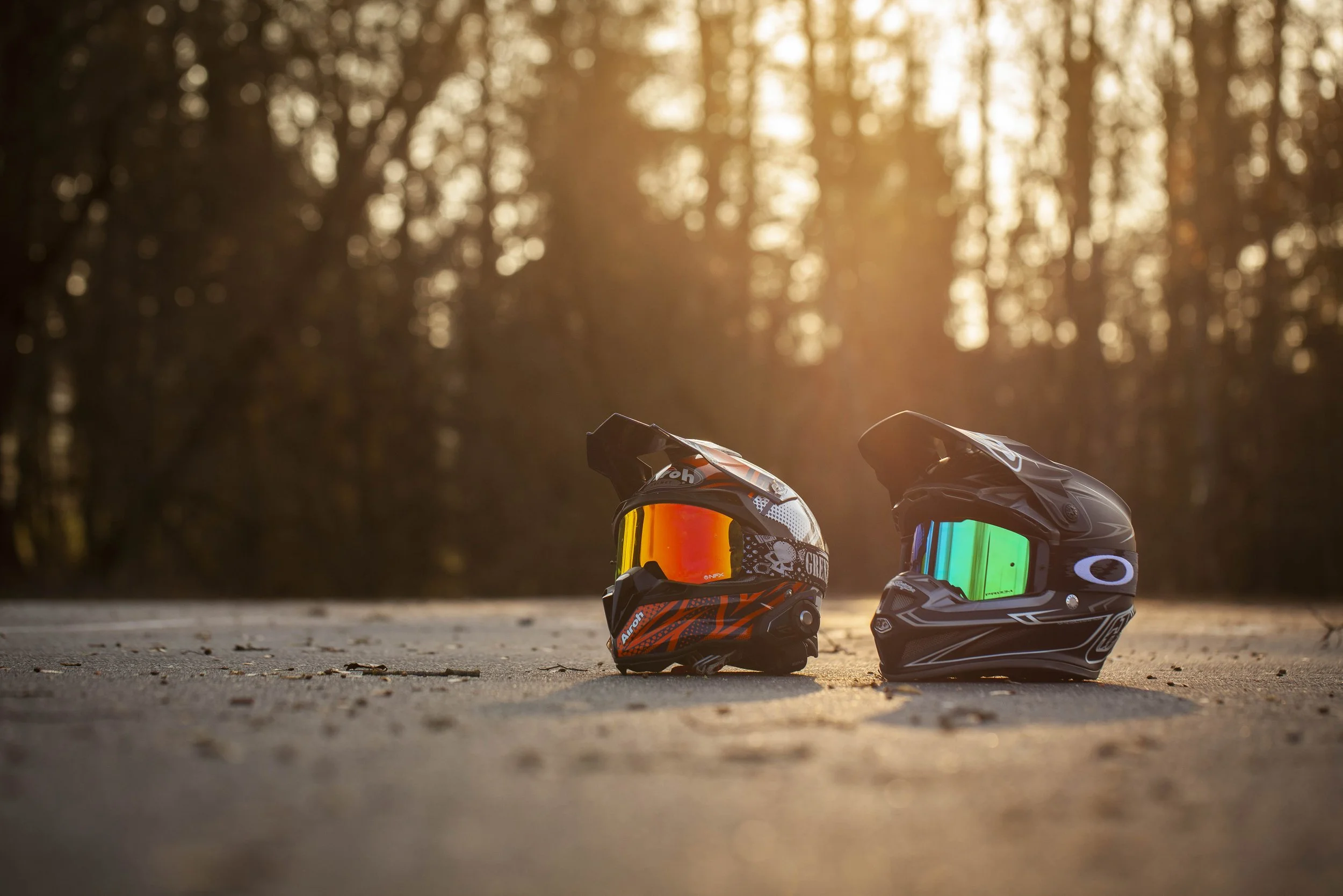 Zwei Motorradhelme im Freien bei Sonnenuntergang, auf einer Asphaltfläche stehend, mit Bäumen im Hintergrund.