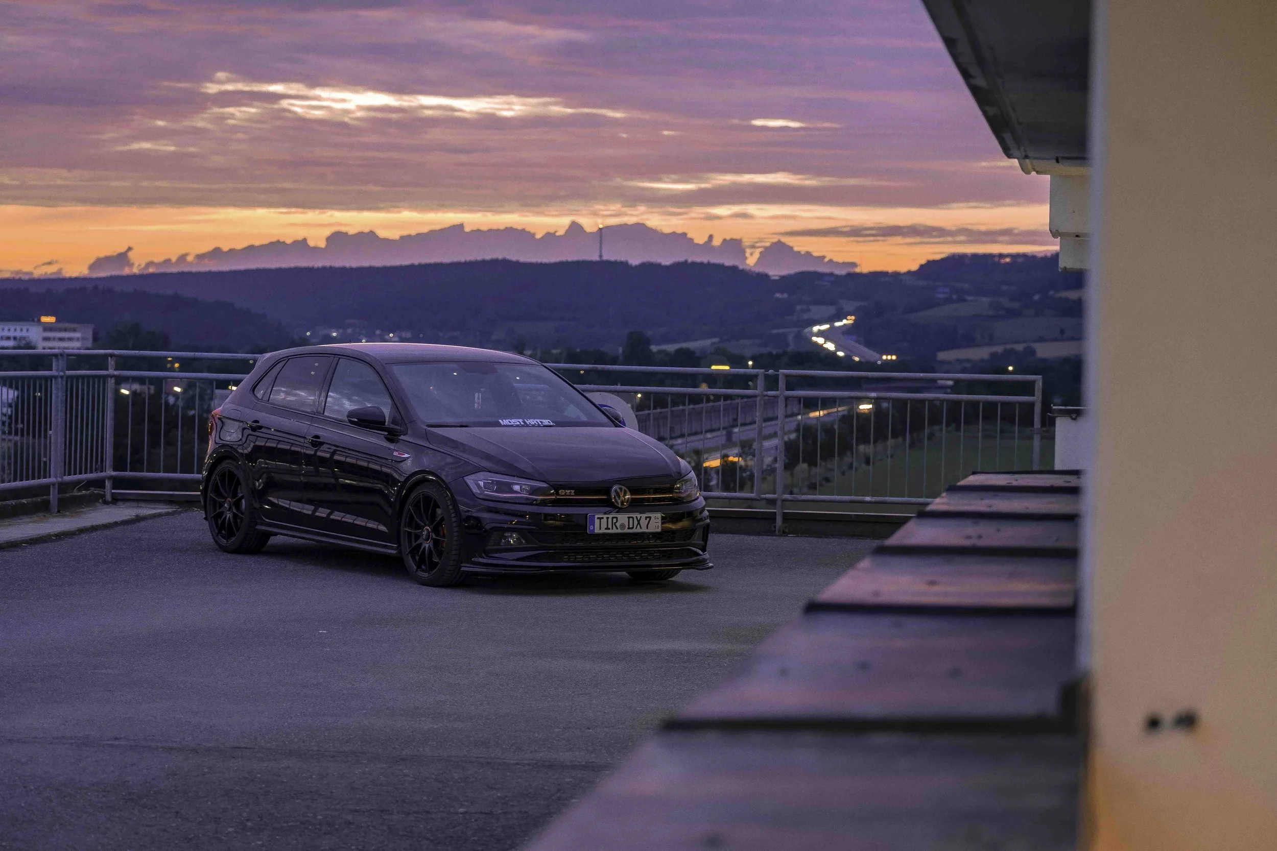 Schwarzer VW Polo GTI steht auf einem Parkplatz mit einem schönen Sonnenuntergang im Hintergrund, umgeben von ländlicher Landschaft und beleuchteter Straße in der Ferne.
