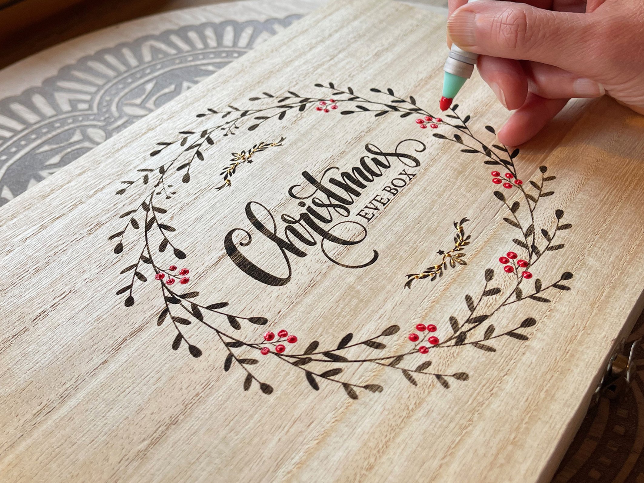 Person decorating a wooden Christmas Eve box with a wreath design and berries.