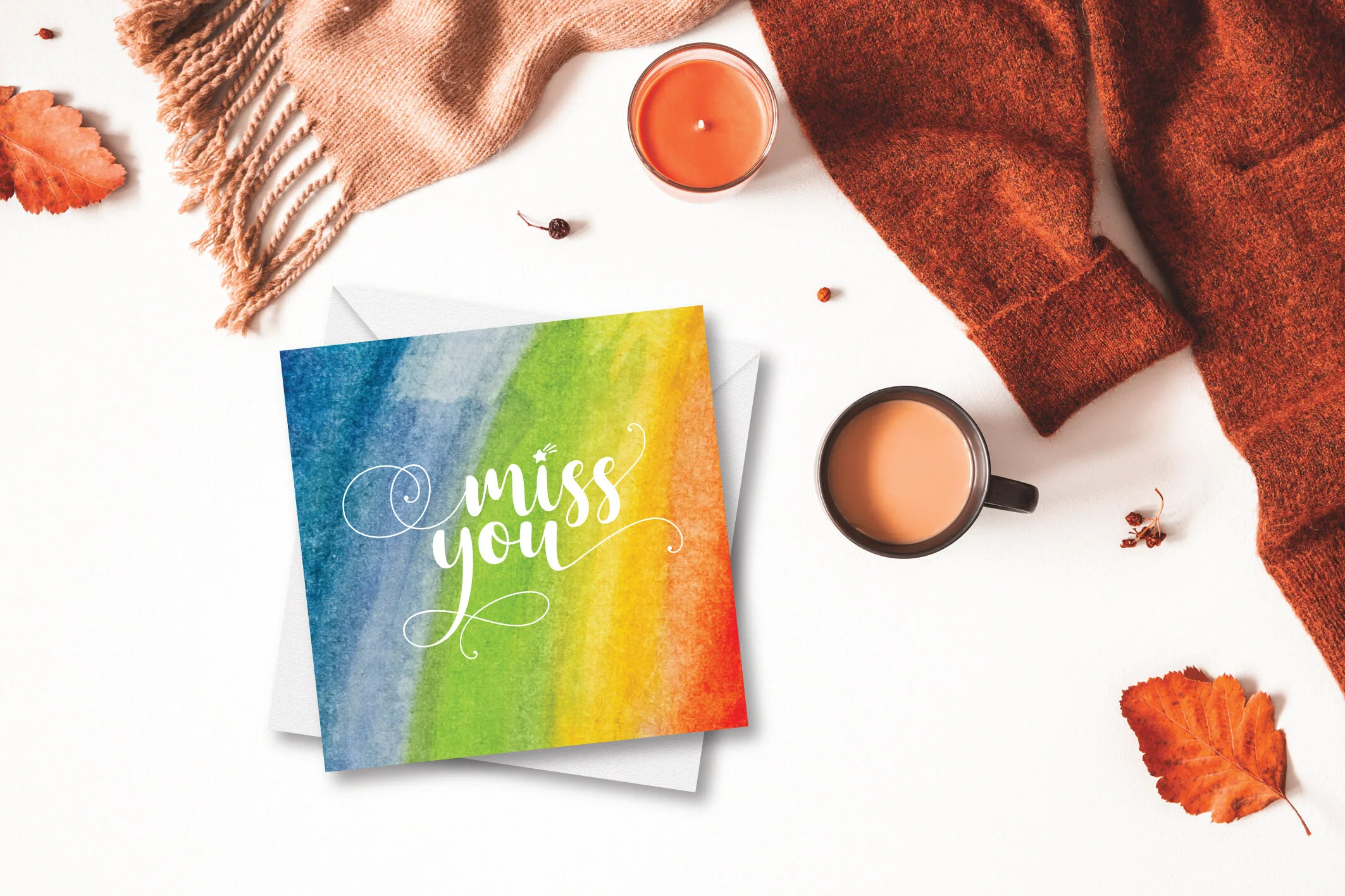 Fall-themed flat lay with a "miss you" card, a warm beverage in a black mug, a burning orange candle, a brown sweater, a beige scarf, and scattered autumn leaves.