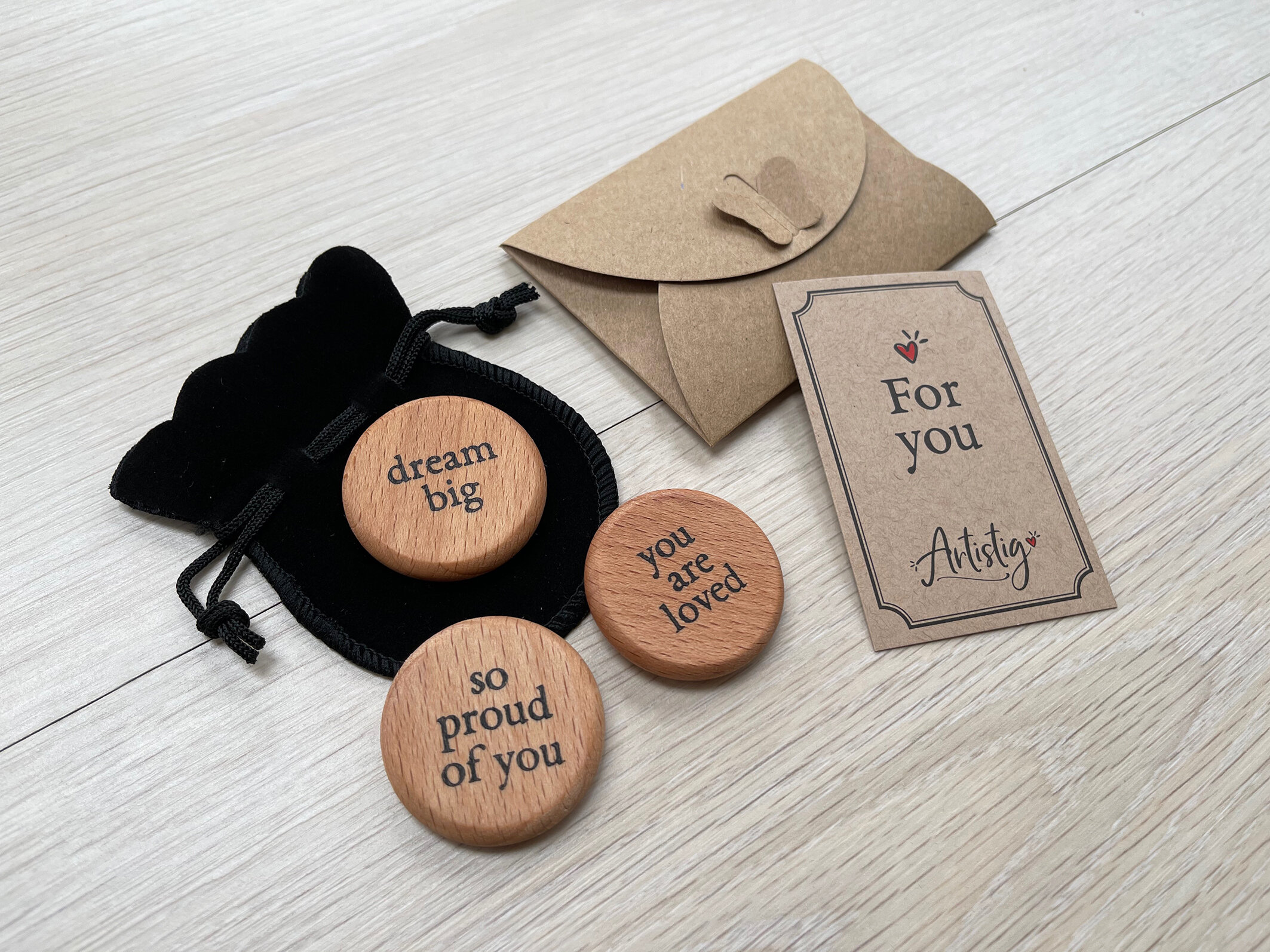 Wooden tokens with inspirational messages "dream big," "so proud of you," "you are loved," a black drawstring pouch, and a card with "For you" alongside a small envelope on a wooden surface.