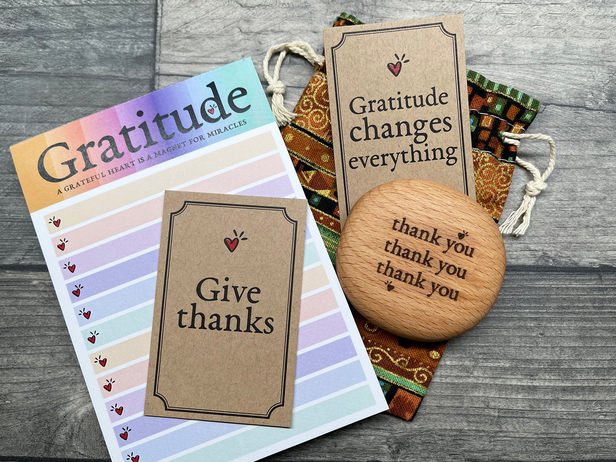Gratitude-themed cards on a wooden surface, including a "Give thanks" card, a "Gratitude changes everything" card, and a wooden token with "thank you" repeated. A colorful gratitude list with spaces to write is also present.