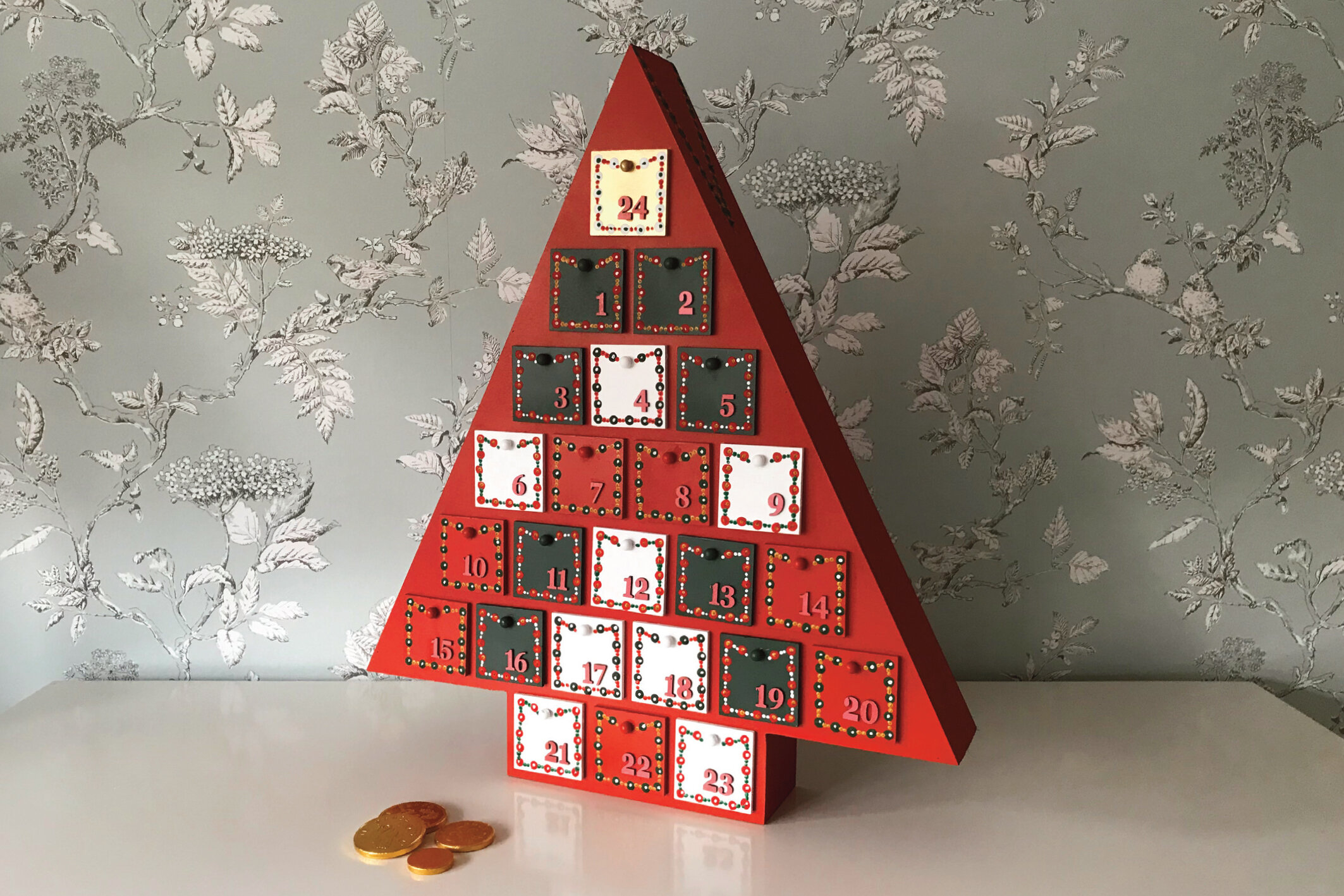 Christmas tree-shaped advent calendar with numbered doors, set against floral wallpaper background, with chocolate coins on the table.