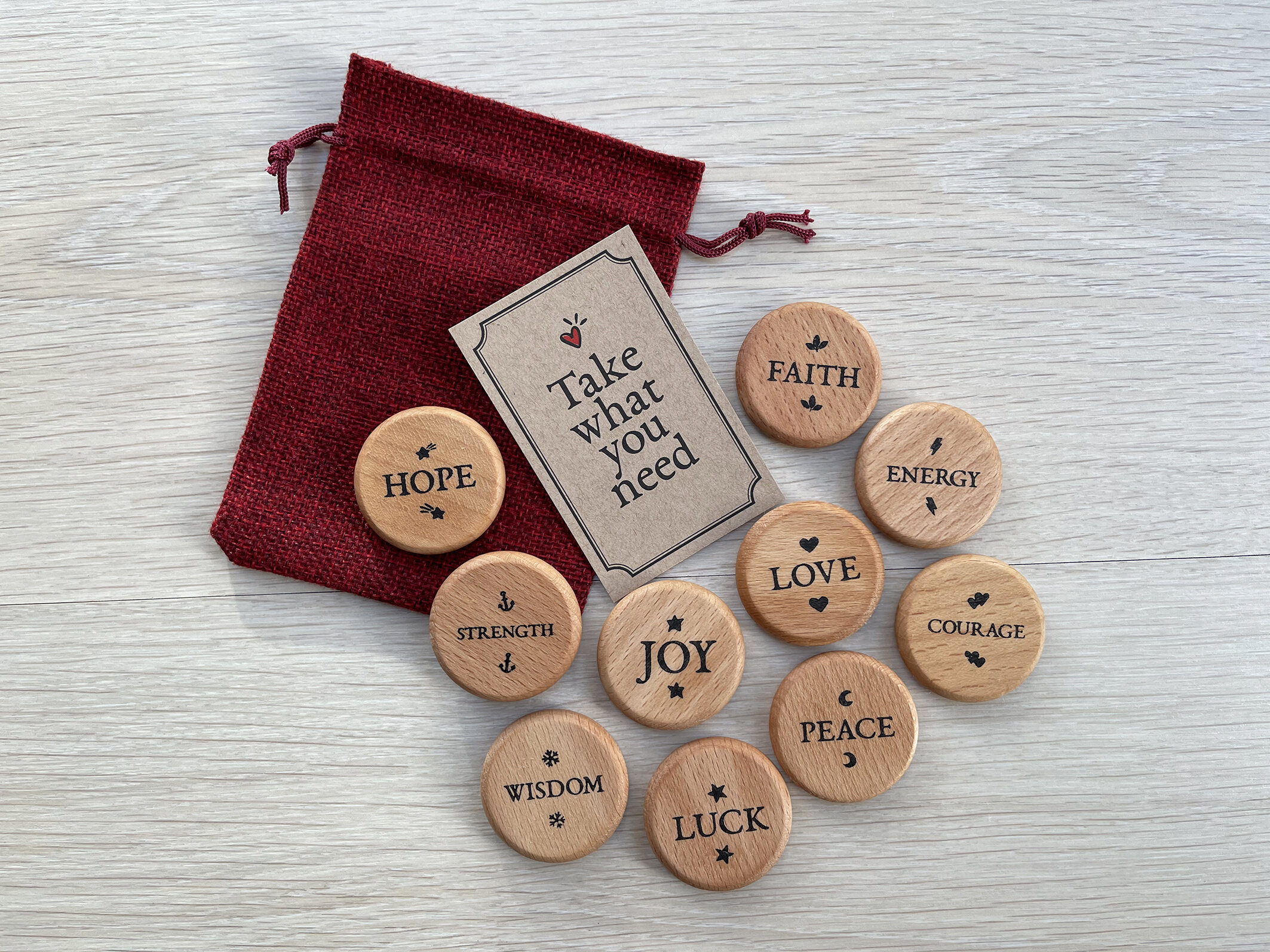 A red drawstring pouch with wooden tokens spread out on a light wooden surface, each inscribed with words like hope, faith, energy, strength, joy, love, courage, wisdom, peace, and luck. A card with "Take what you need" accompanies them.