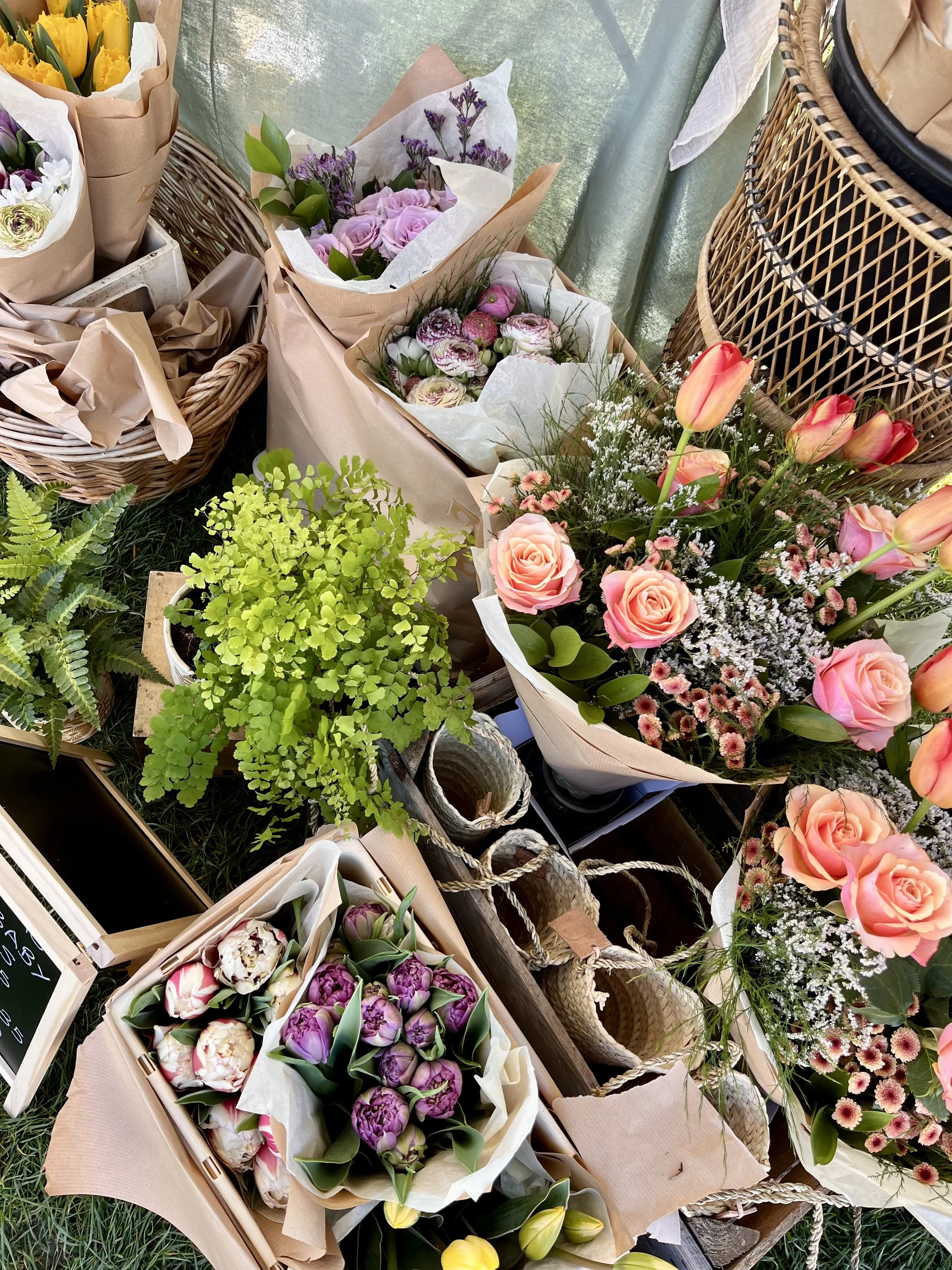 Market flowers using colorful flower bouquets including pink roses, tulips, purple tulips, white flowers, and green foliage, arranged in paper wrapping, baskets, and containers at a market stand.