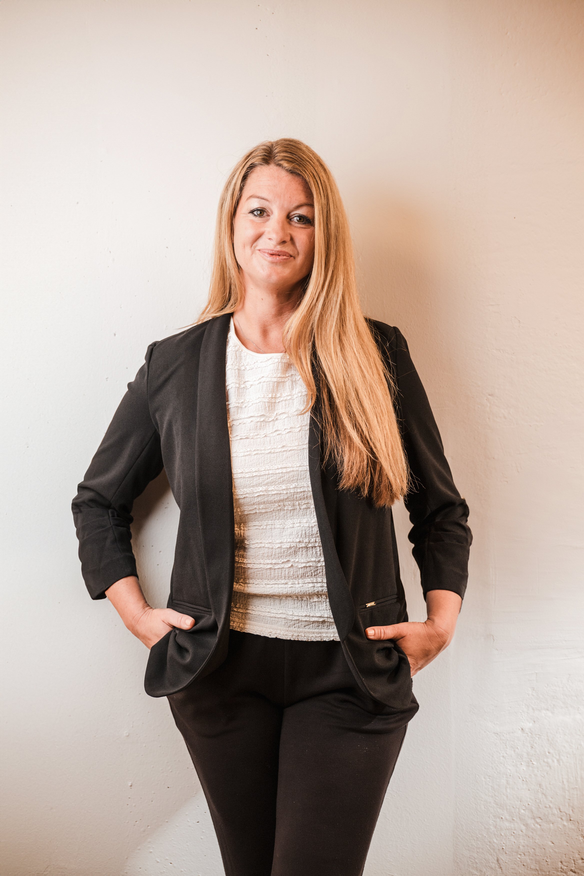 Eine Frau mit langen blonden Haaren trägt einen schwarzen Blazer und eine weiße Bluse, steht vor einer weißen Wand, lächelt leicht und hat beide Hände in den Taschen.