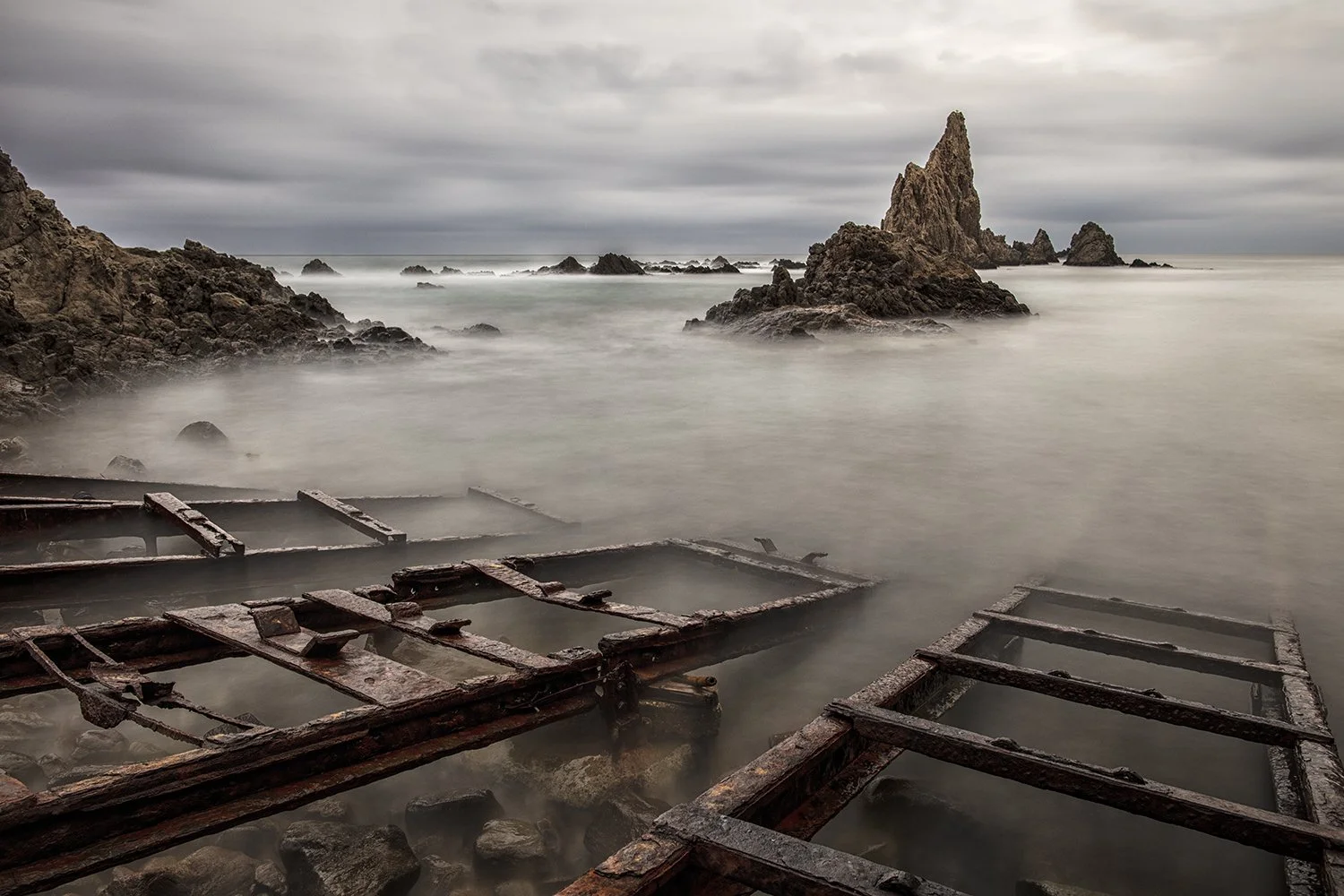 Rusted shipwreck remains on rocky shoreline with foggy ocean and jagged sea stacks under cloudy sky.