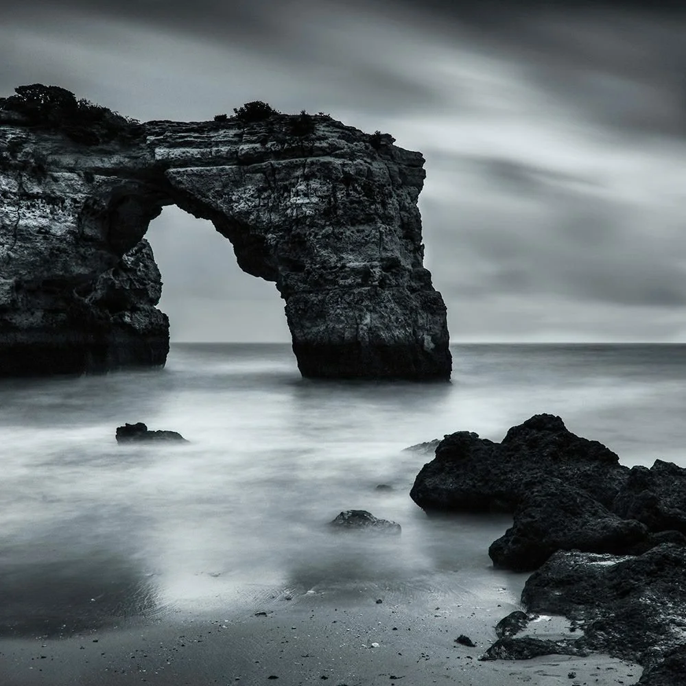 A black and white photo of a natural rock formation with a large arch over the ocean, with rocky shoreline in the foreground and a cloudy sky overhead.