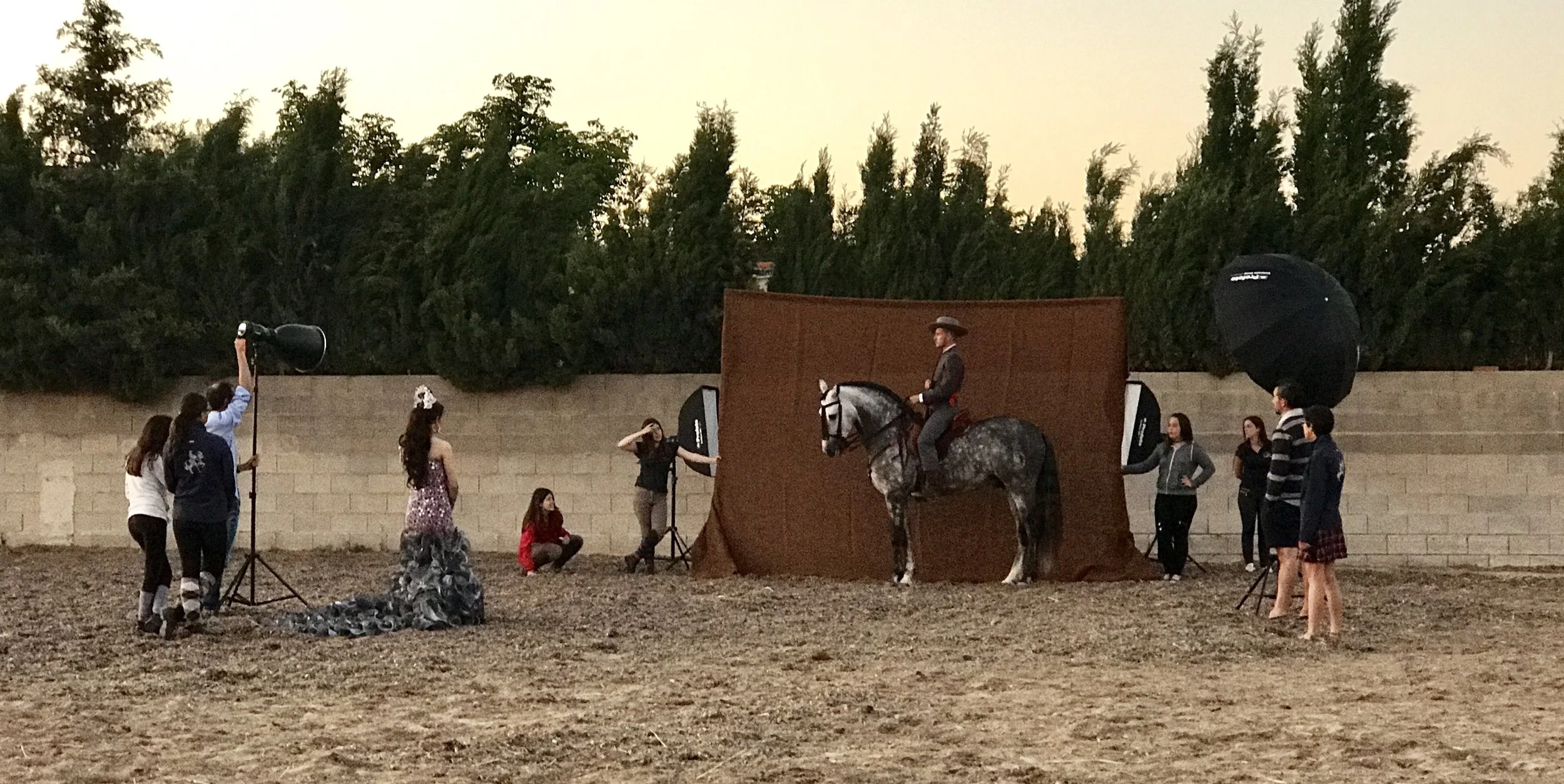 Photoshoot with a man on horseback in front of a brown backdrop, surrounded by Didier Borgeaud  and crew members using lighting equipment outdoors, with a brick wall and trees in the background.