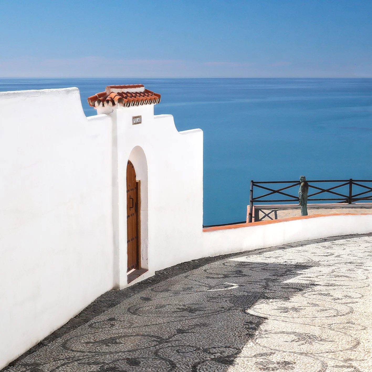 White Mediterranean-style building with a wooden door, red-tiled roof, and a sign reading 'Pilar,' overlooking a blue ocean.
