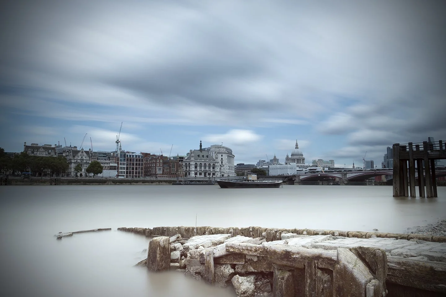 Long exposure photograph of the London skyline across the River Thames, with clouds moving over historic and modern buildings, including St. Paul's Cathedral, and an old pier in the foreground.