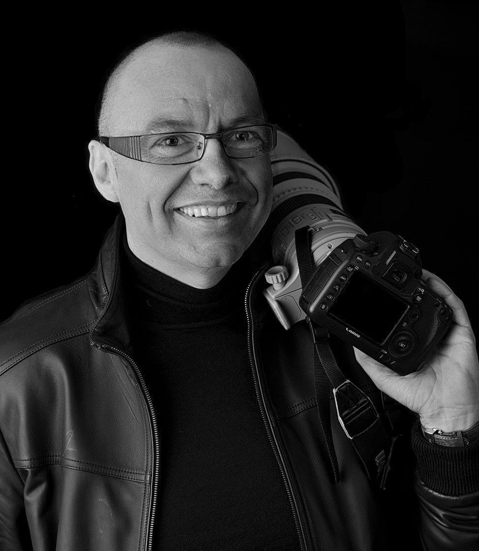 Didier Borgeaud smiling with his glasses and wearing a leather jacket, holding a professional camera with a large zoom lens against a black background.