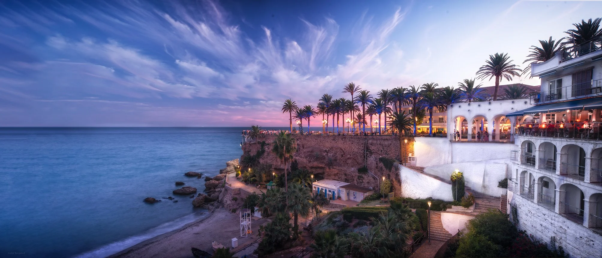 Sunset view of a coastal hotel with palm trees, outdoor dining area, and rocky shoreline.