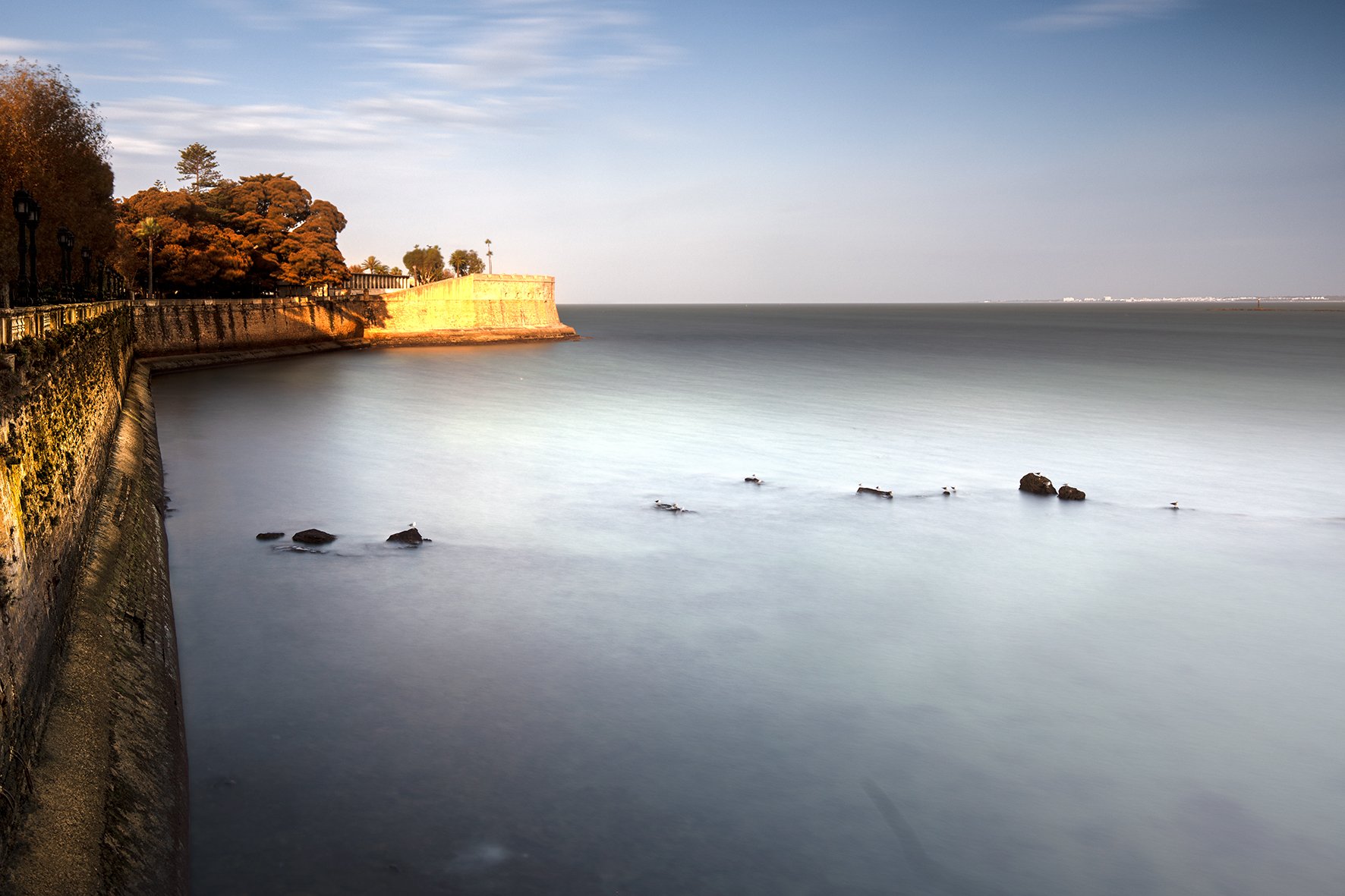 Coastal scene with a stone seawall on the left, trees with autumn foliage, calm water, and a distant horizon under a partly cloudy sky.