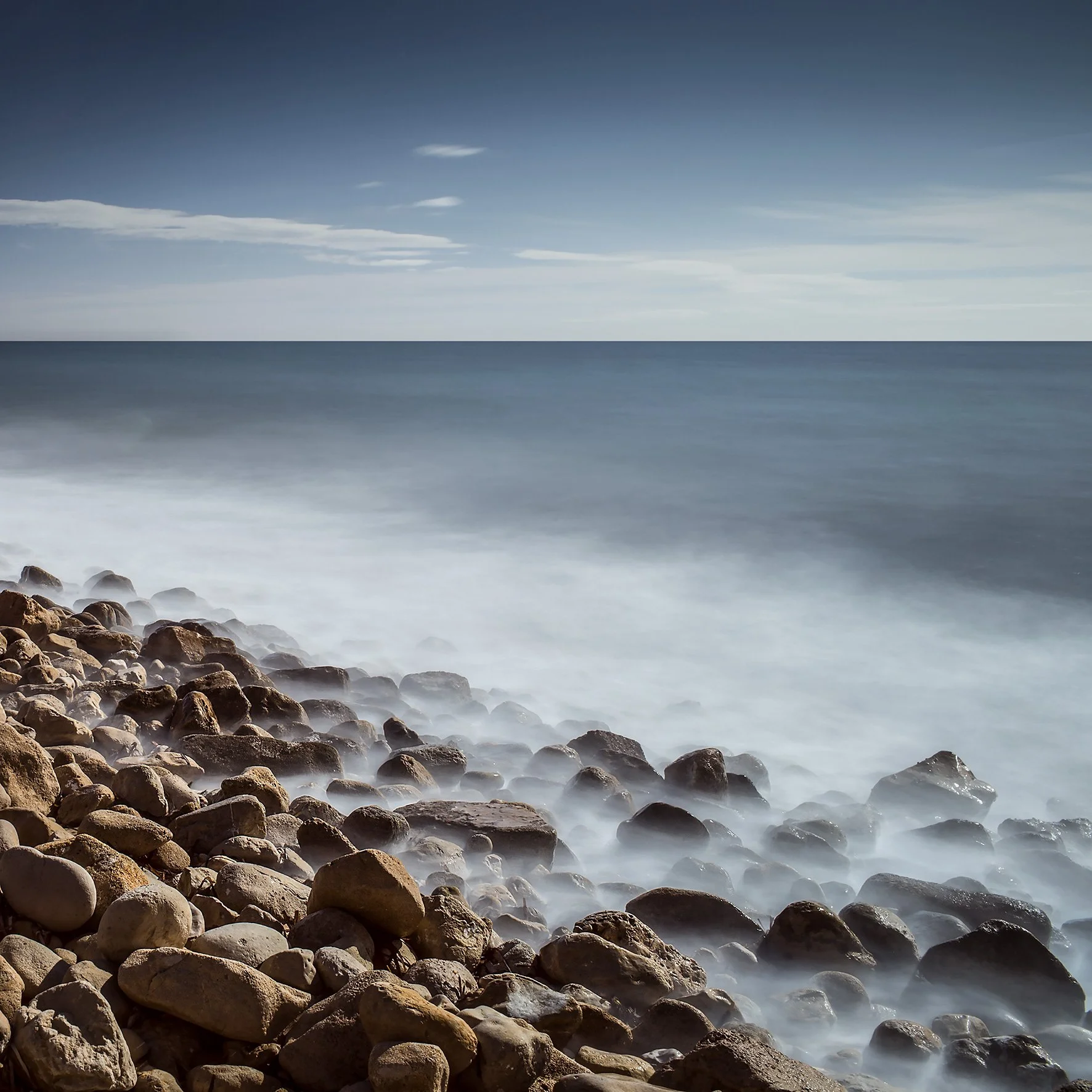 Long exposure photo of a rocky shoreline with blurred ocean waves under a cloudy sky.