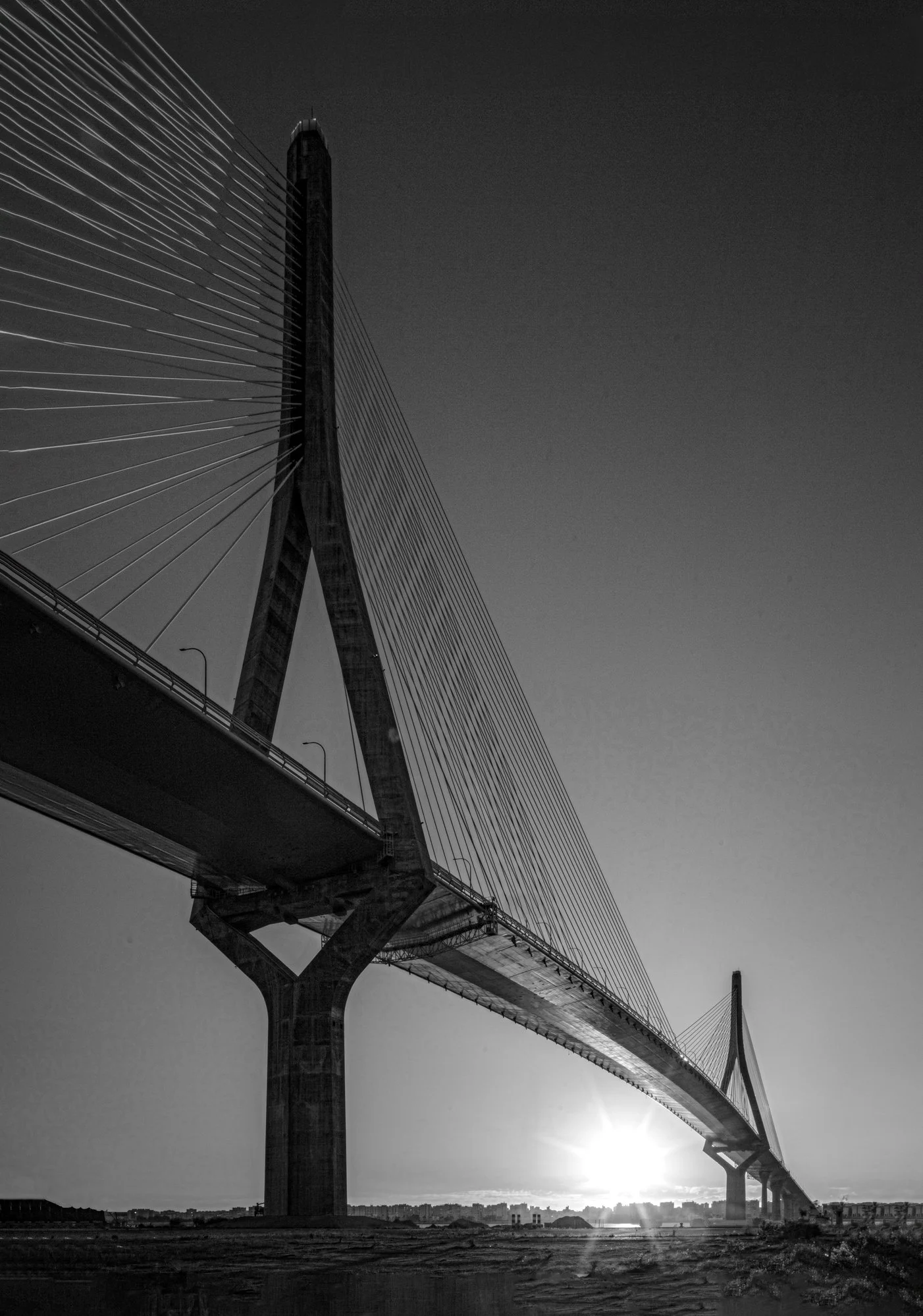 Black and white photo of a modern cable-stayed bridge with a tall pylon and numerous cables extending to the deck, with the sun near the horizon.