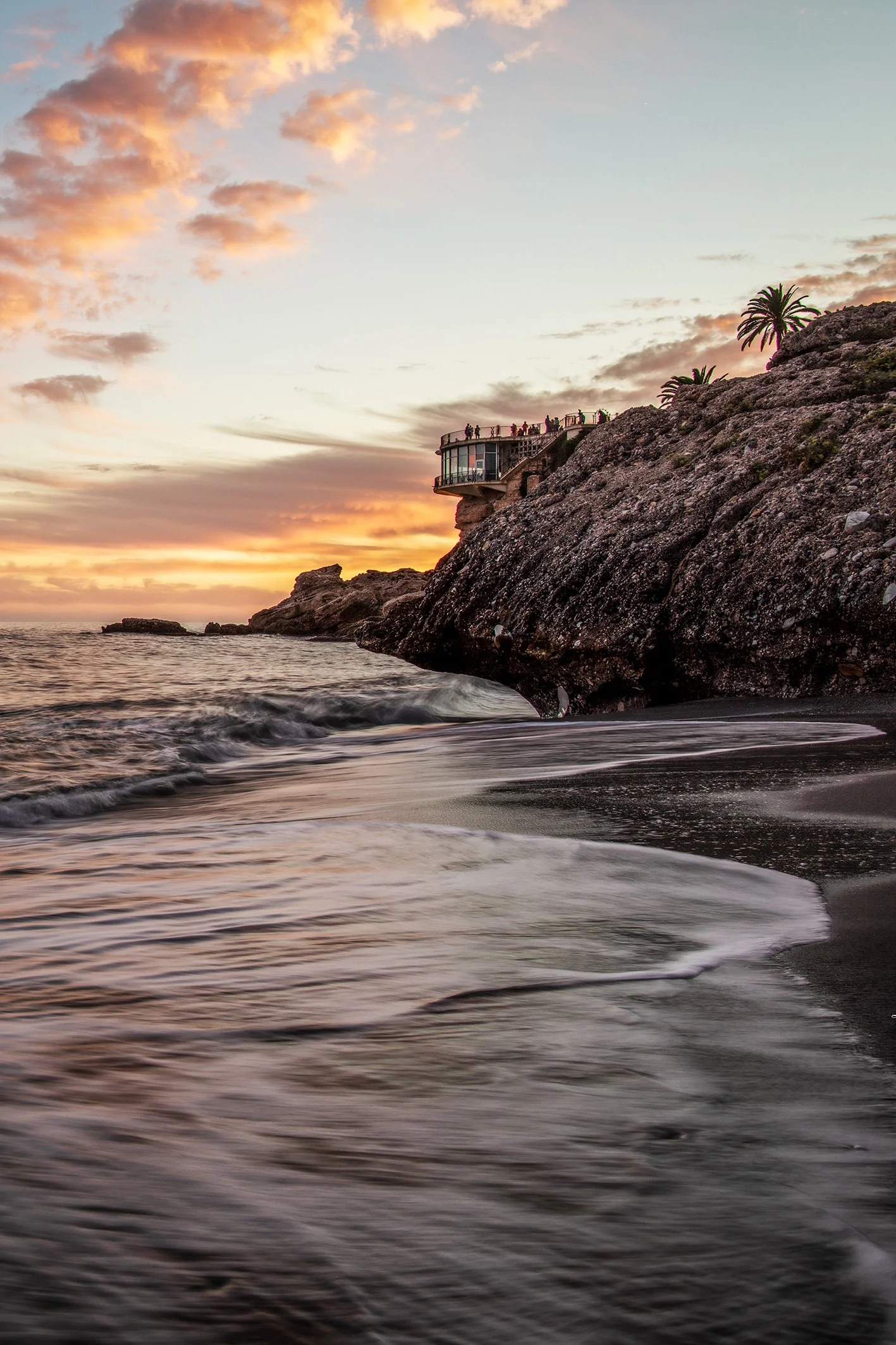 A scenic beach scene at sunset with waves gently washing onto the dark sandy shore, large rocky formations, a cliff with a curved viewing platform, and a few palm trees in the background.
