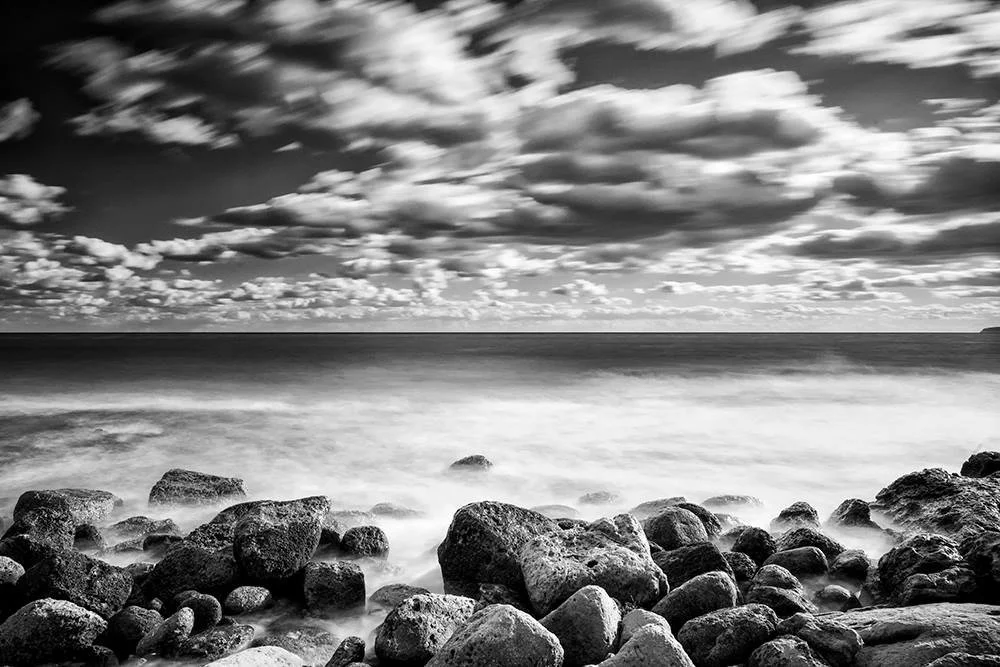 Black and white photo of a rocky coastline with clouds in the sky and the ocean in the background.