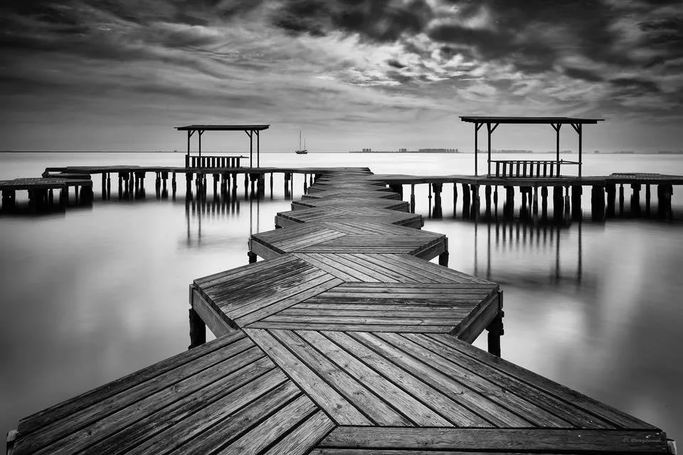 A black and white photo of a wooden pier extending over calm water with two small covered sitting areas at the end, under a cloudy sky in the distance.