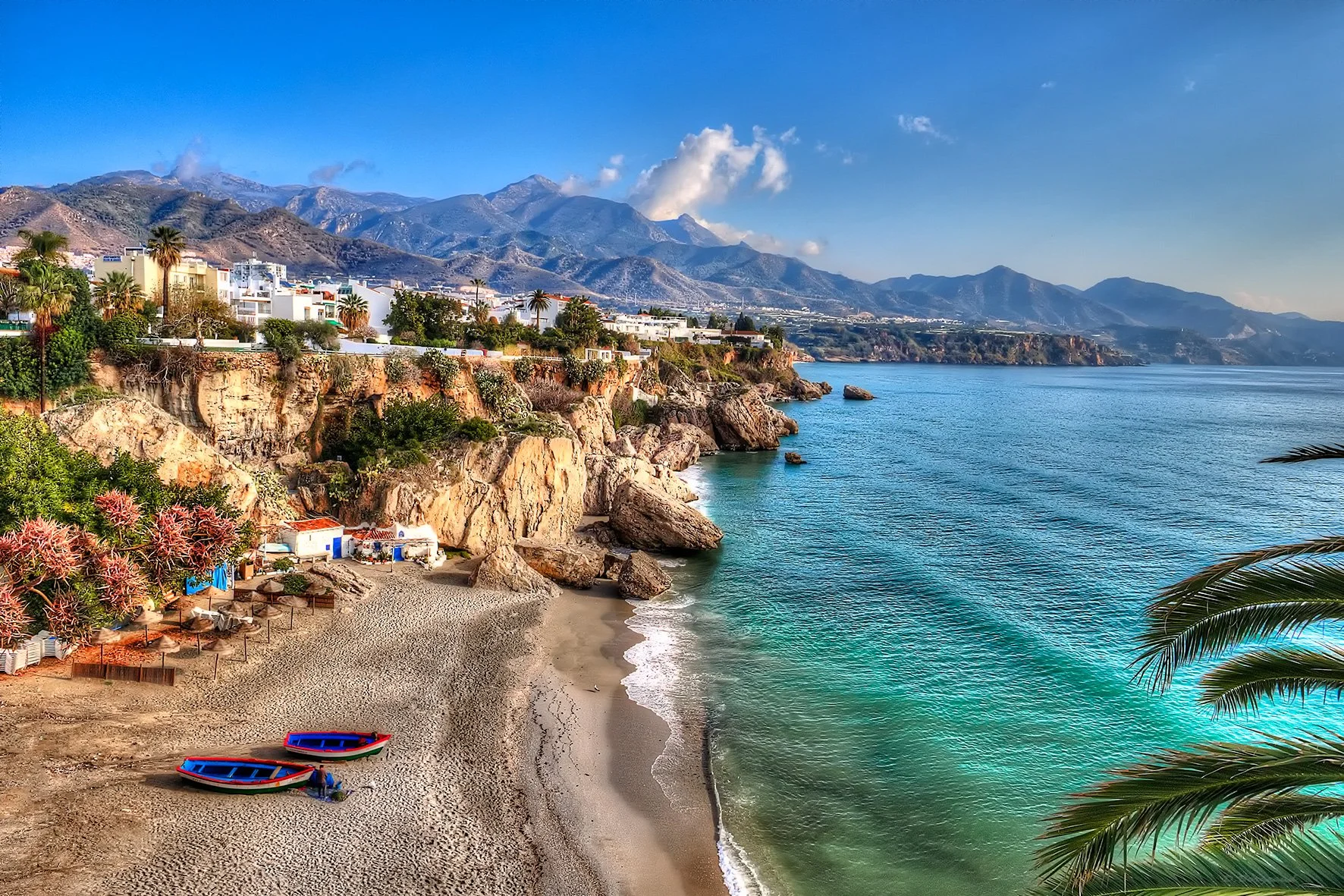 A scenic coastal landscape with sandy beach, turquoise water, rocky cliffs, palm trees, beach chairs, and mountains in the background under a clear blue sky.