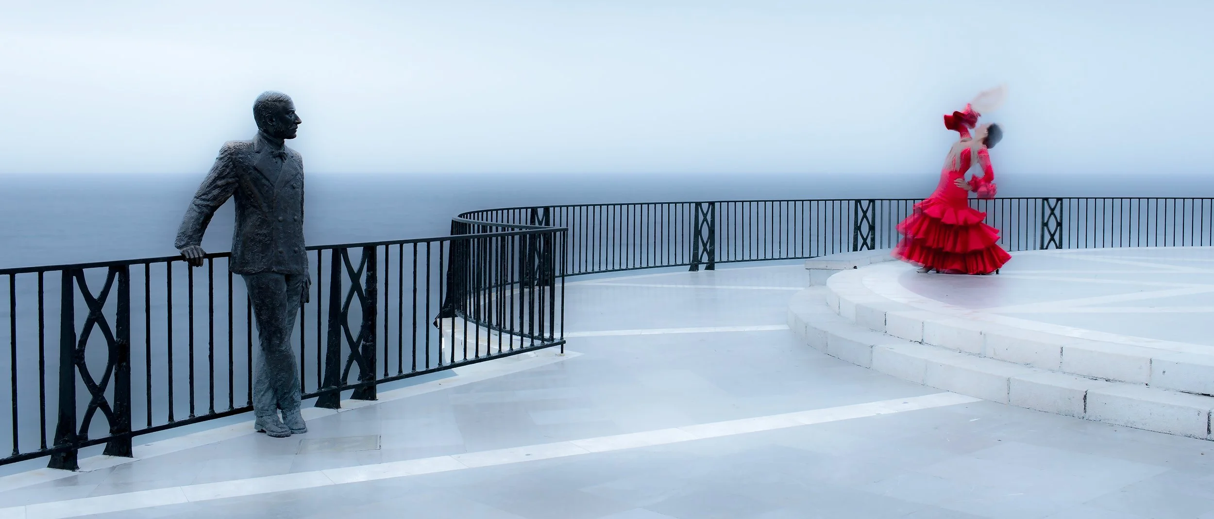 Statue of a man in a suit leaning against a black railing and a woman in a red flamenco dress standing on white steps, overlooking the ocean under a cloudy sky.