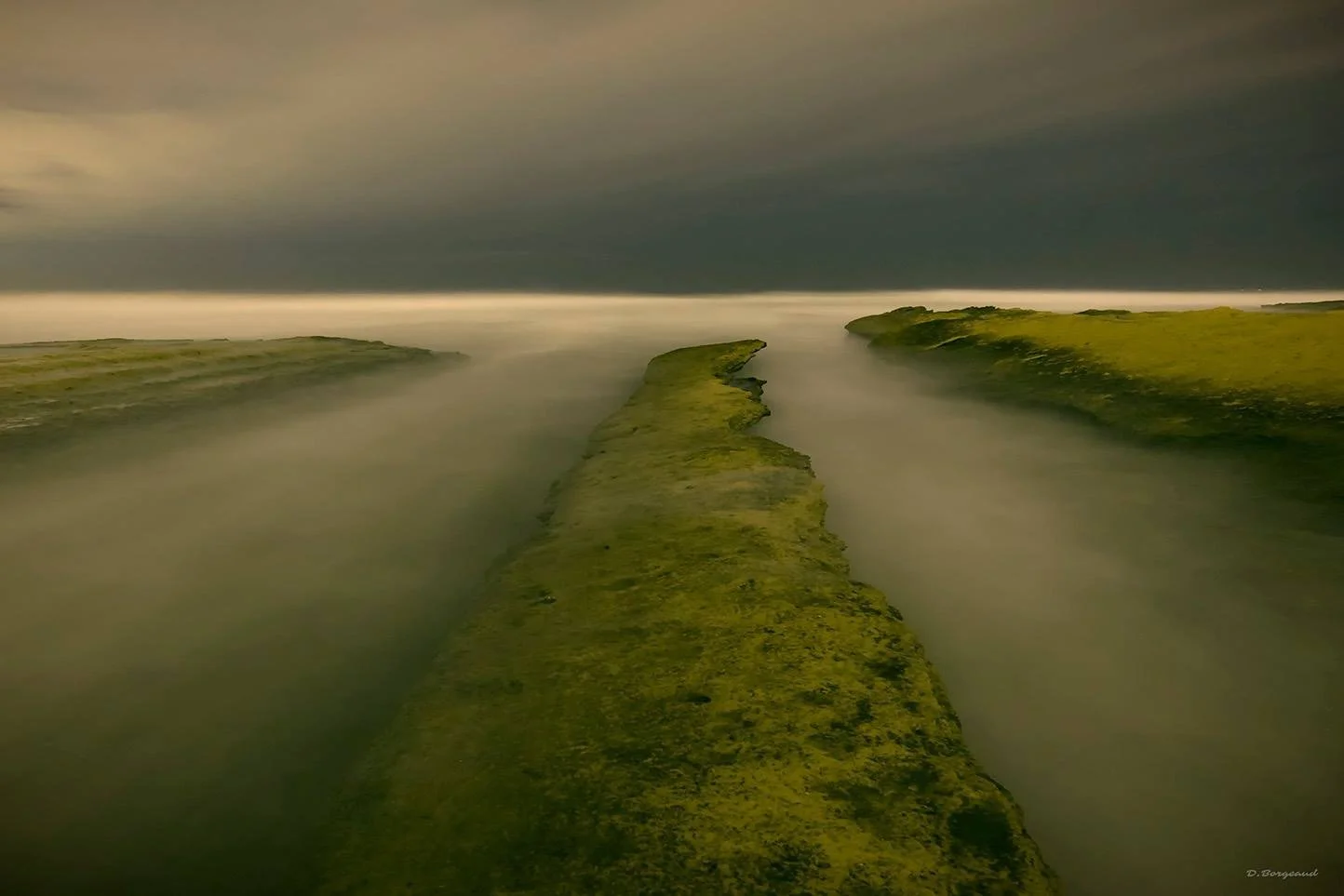 Long moss-covered rock formation extending into the ocean under a dark stormy sky with blurred water and clouds.