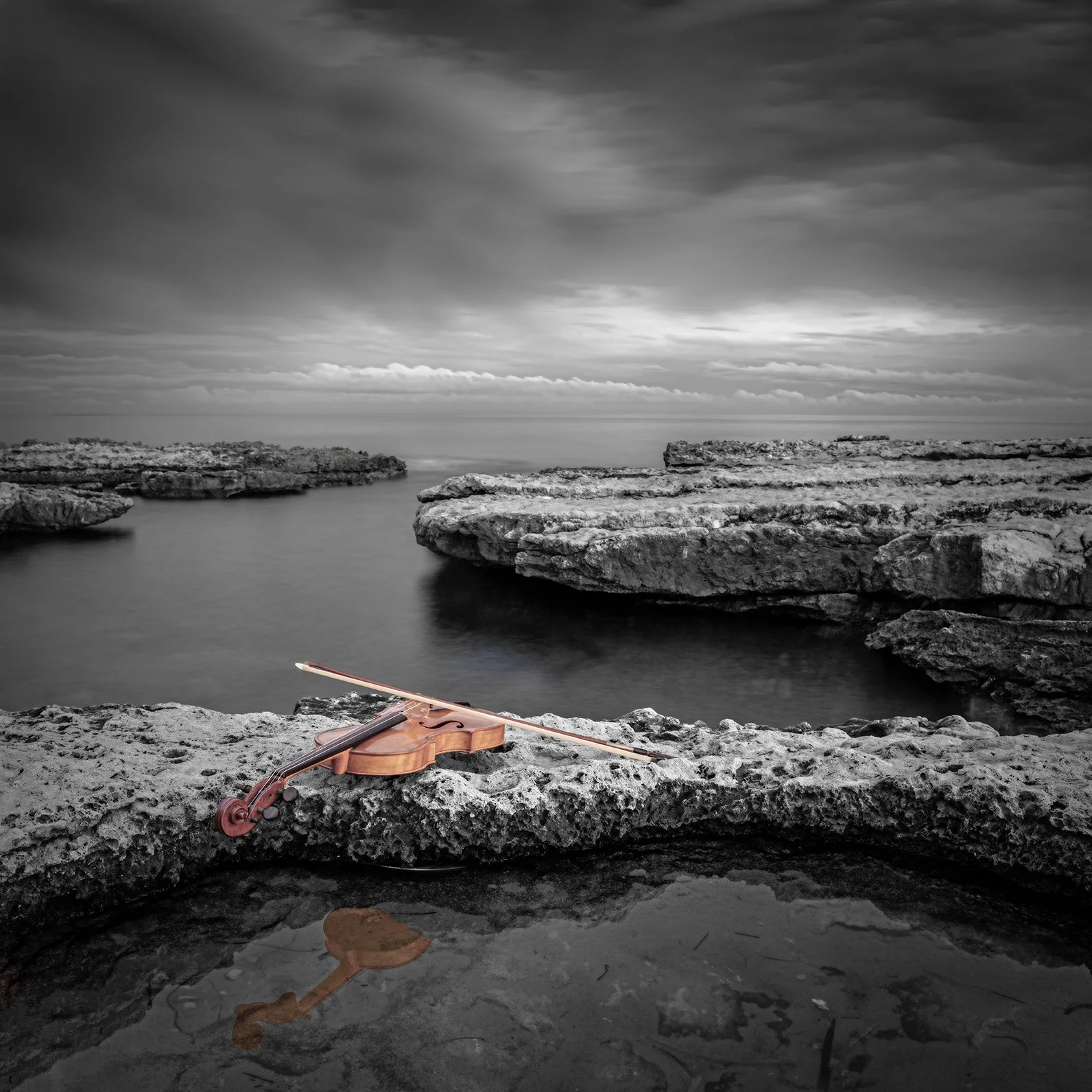 A violin with a bow resting on a rocky shoreline with water and cliffs in the background, black and white photo.