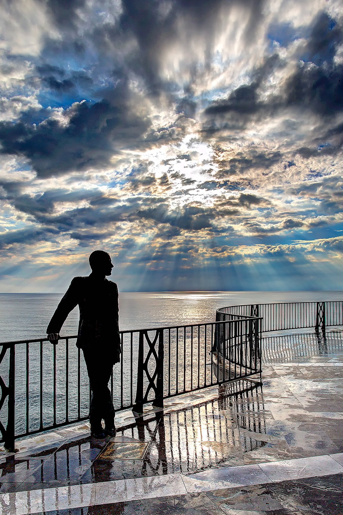 Silhouette of a person standing near a railing on a terrace overlooking the ocean, with dramatic cloud-filled sky and rays of sunlight breaking through.