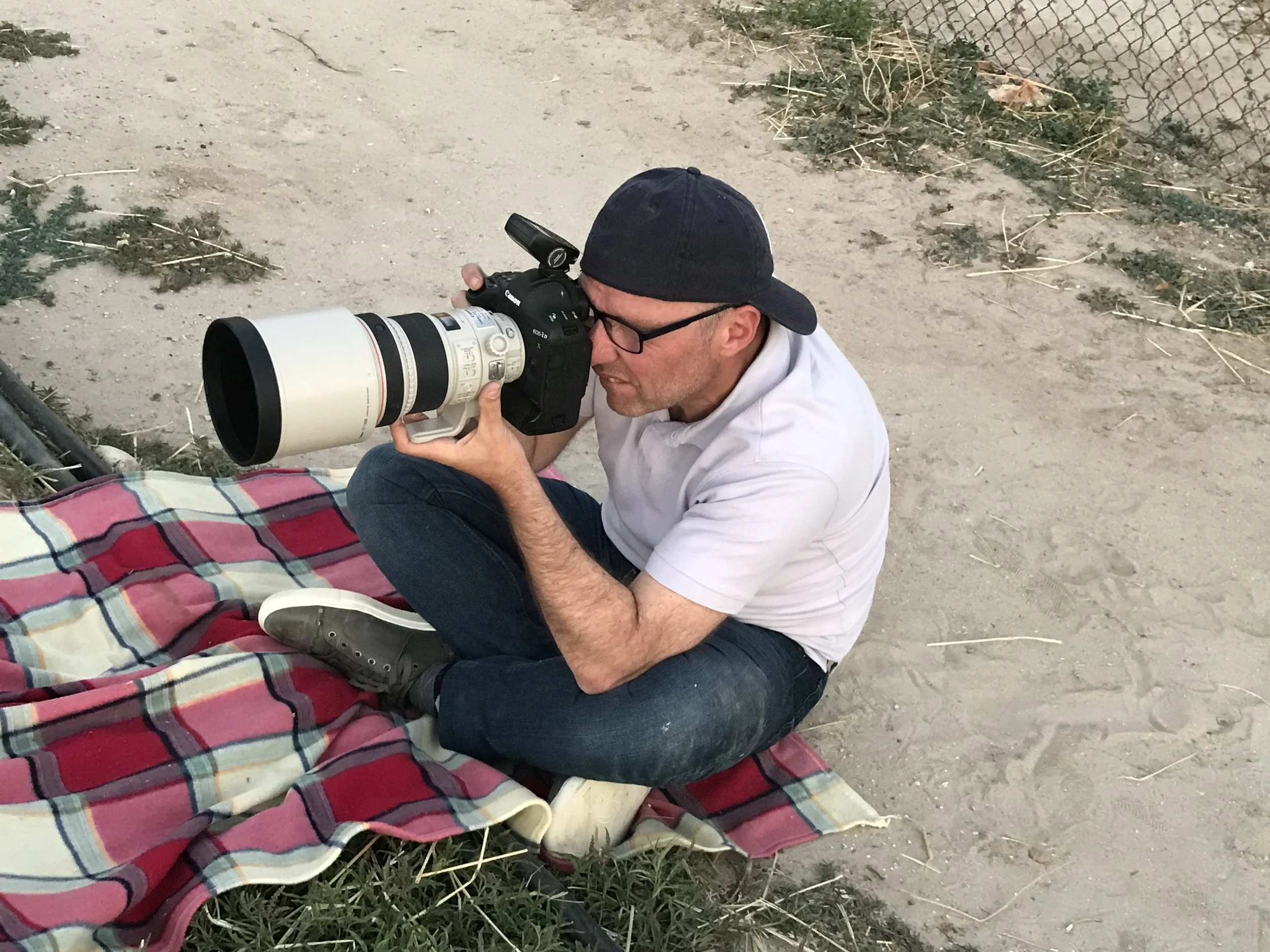 Didier Borgeaud  sitting on the ground with crossed legs, wearing glasses, a black cap backwards, white shirt, and jeans, taking a photograph with a large professional camera.