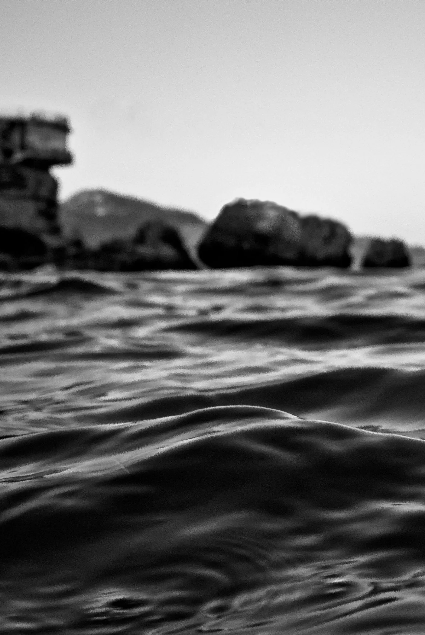 A black and white photo showing the surface of the water in the foreground and rocks and a building in the distance on the shoreline.