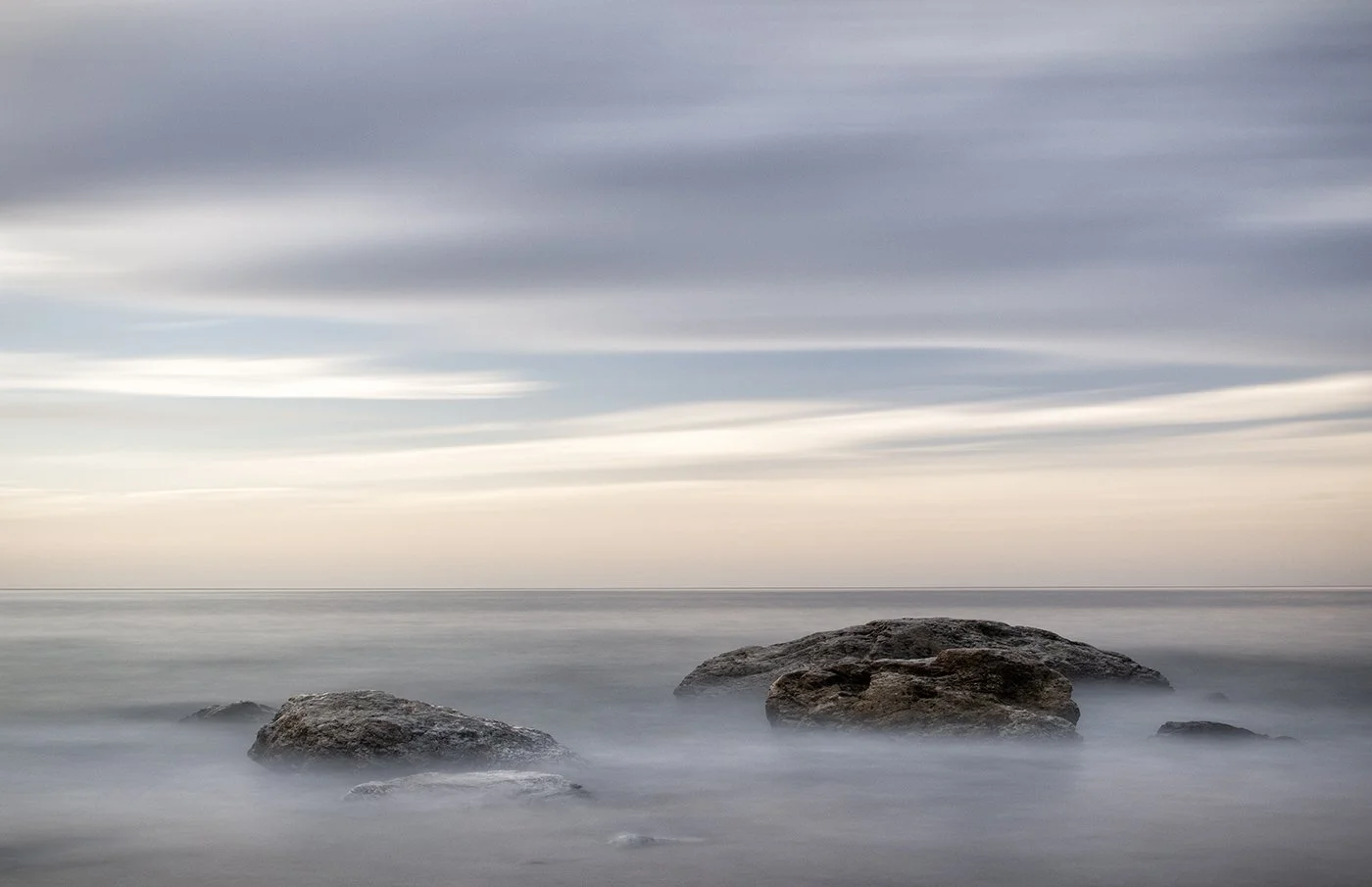 Three large rocks in calm ocean water under a cloudy sky at sunset or sunrise.