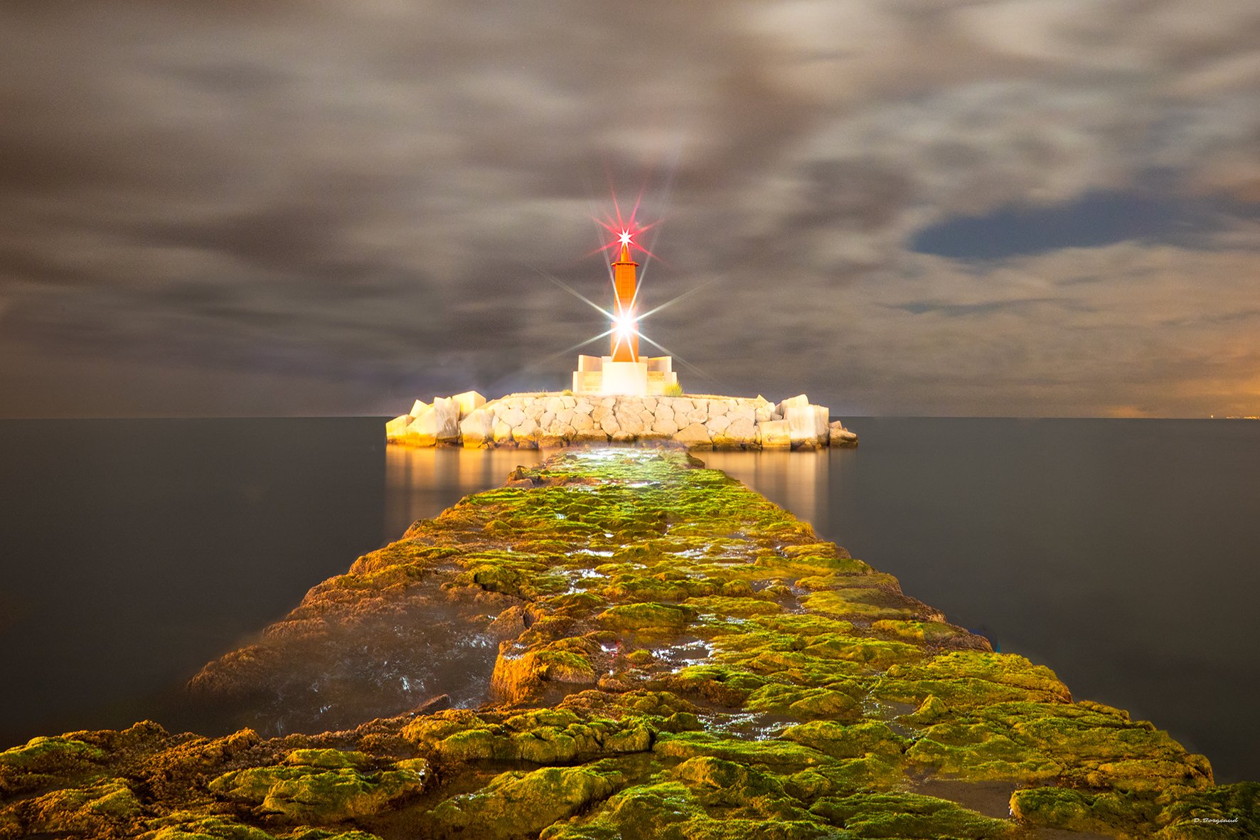 A rocky moss-covered pier extending into the water leads to a lighthouse on a small rocky island, with cloudy sky and distant lights in the background at night.