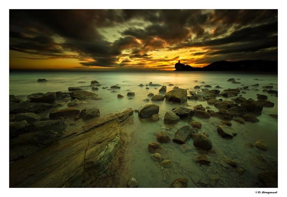 Rocks along a rocky shoreline during sunset with dark clouds and a silhouette of a small island or structure in the distance.