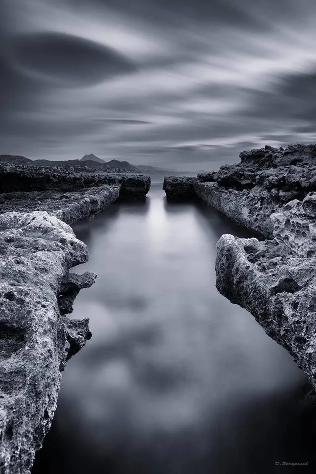 Black and white long exposure photo of a narrow water channel between jagged rocks, with moonlit clouds in the sky and distant mountains on the horizon.