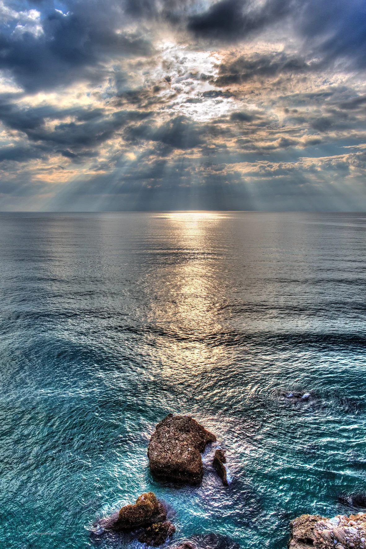A seascape with rocks in the foreground, calm ocean waters, and a dramatic sky with clouds and sun rays shining through.