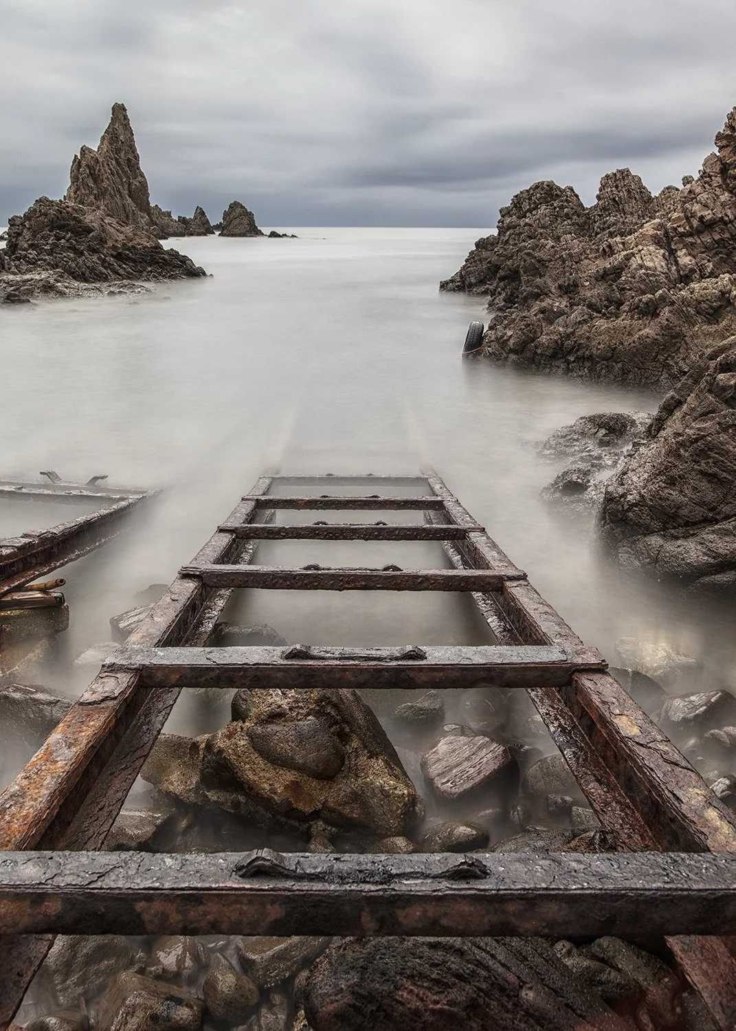 Rusty, partially submerged wooden ladder leading into calm water between rocky cliffs on a cloudy day.