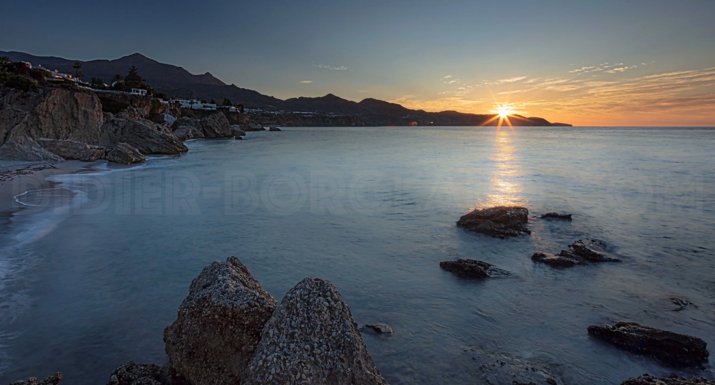 Sunset over a calm ocean with rocky shoreline and distant mountains.