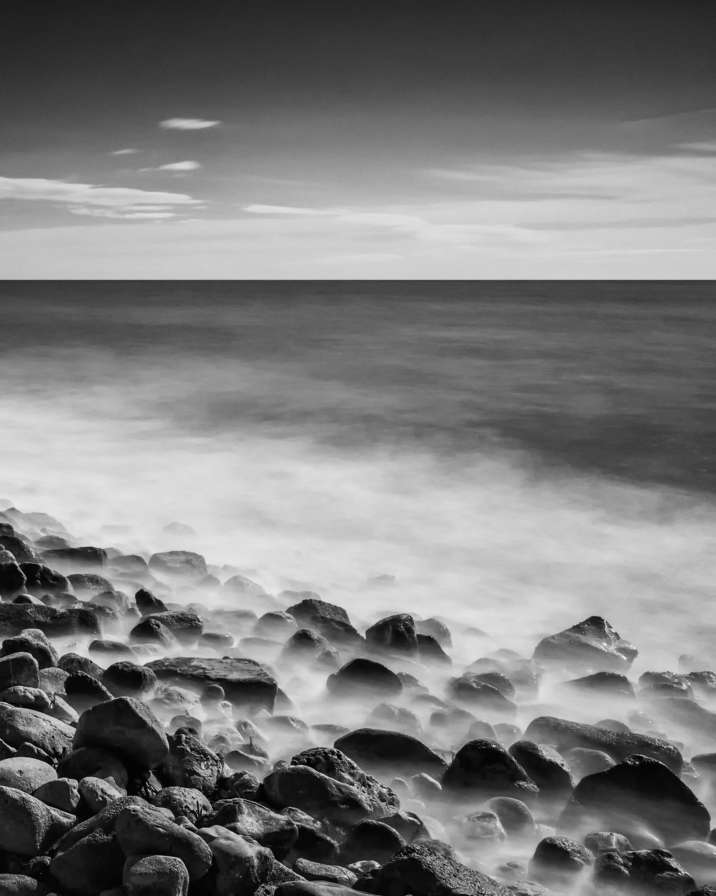Black and white photo of a rocky shoreline with waves washing over the rocks under a cloudy sky.